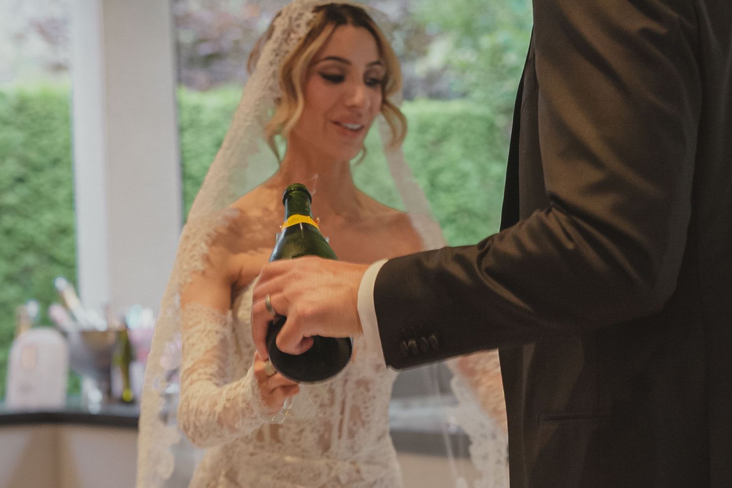 Bride in lace veil and groom in black suit opening champagne bottle at wedding reception.