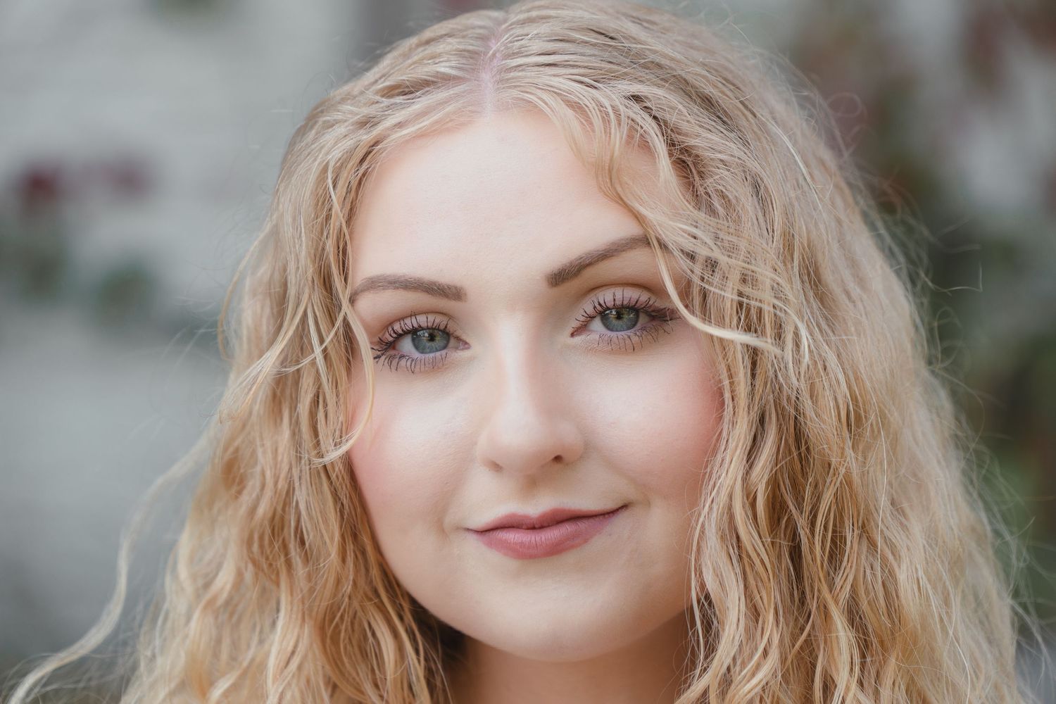 Young woman with curly blonde hair and blue eyes smiling softly in a professional portrait photo.