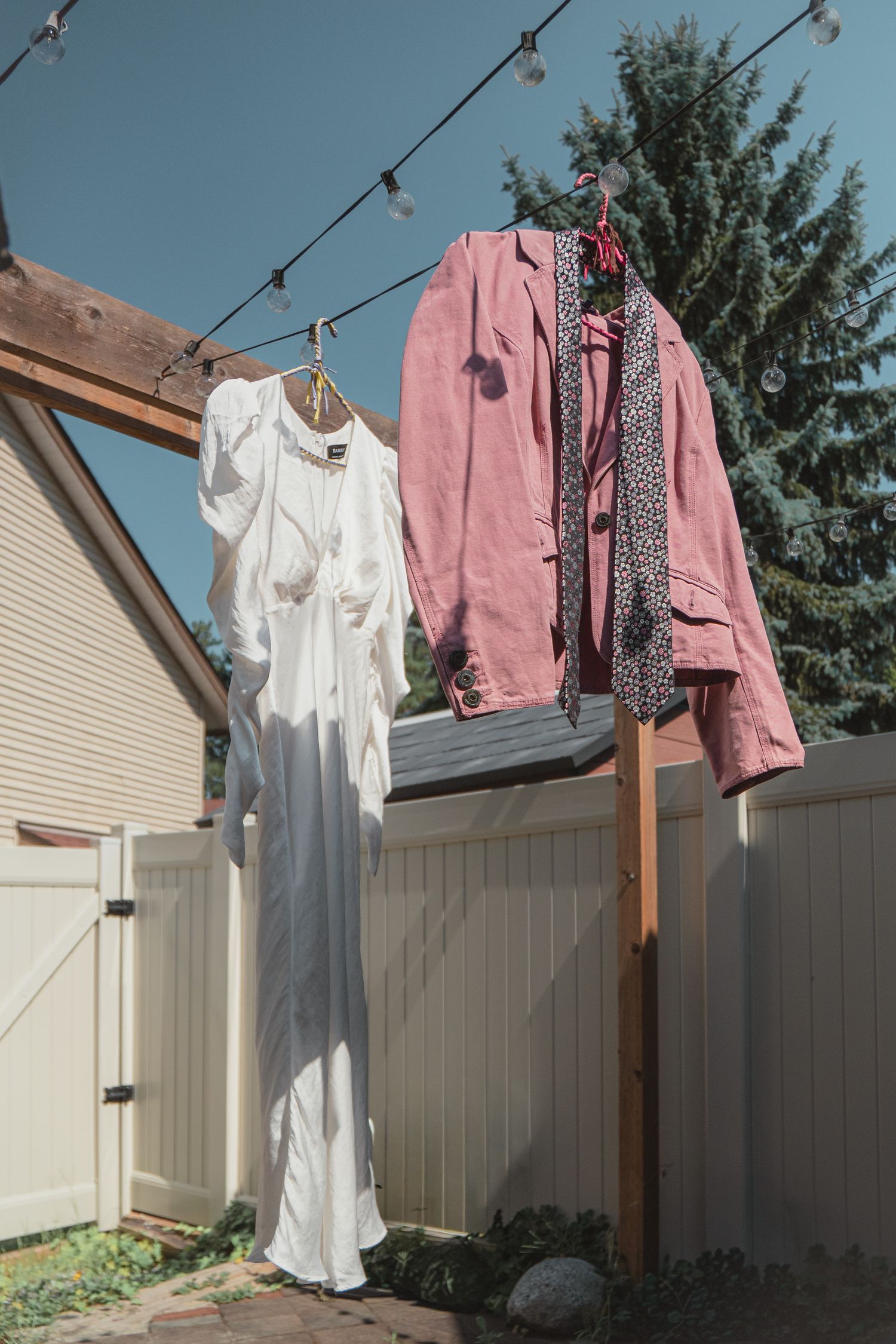 White dress and pink jacket hanging on wooden post with string lights in sunny backyard near white fence.