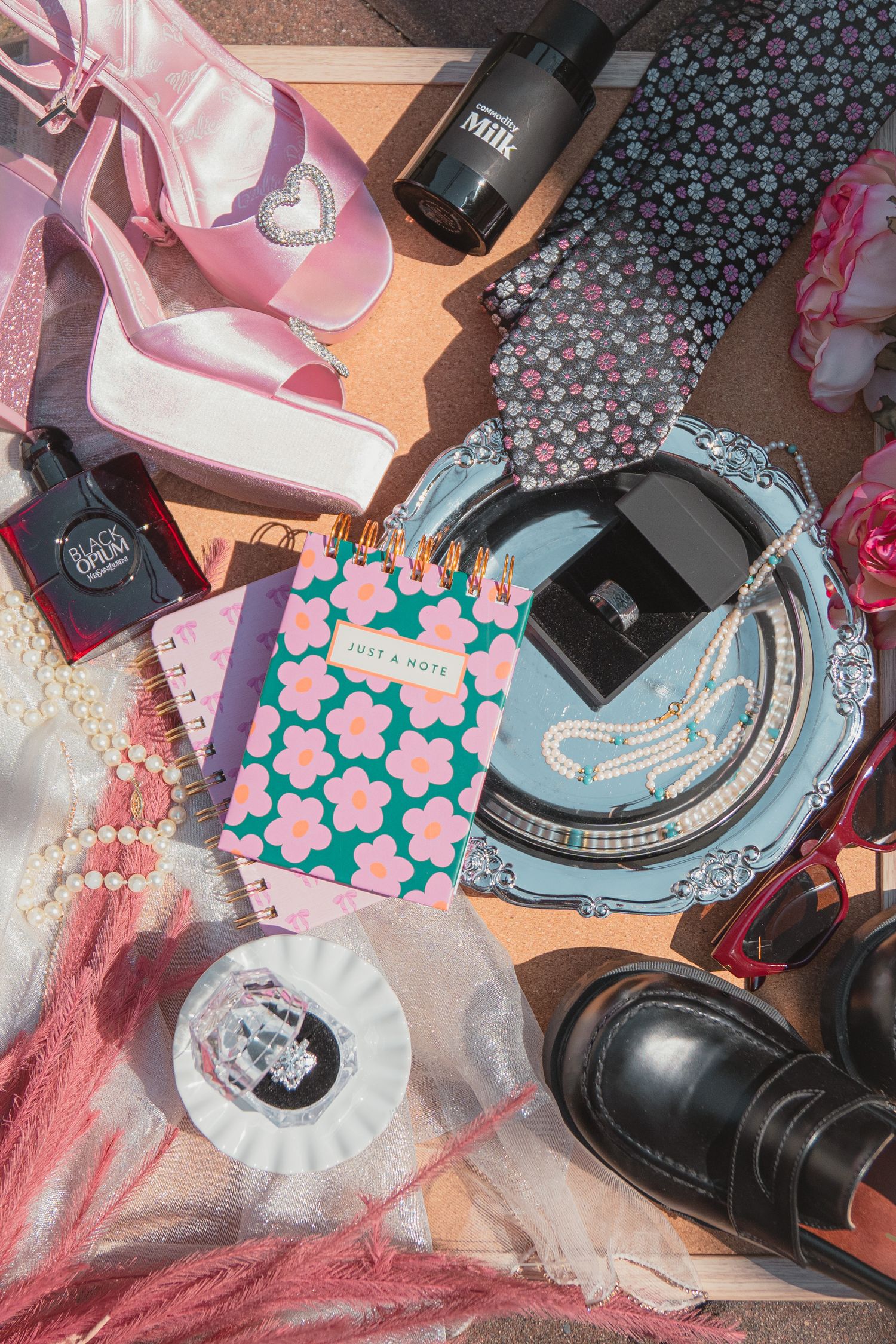 Flat lay of trendy accessories including pink heels, floral notebook, pearl necklace, black boots, and beauty items on cork board.