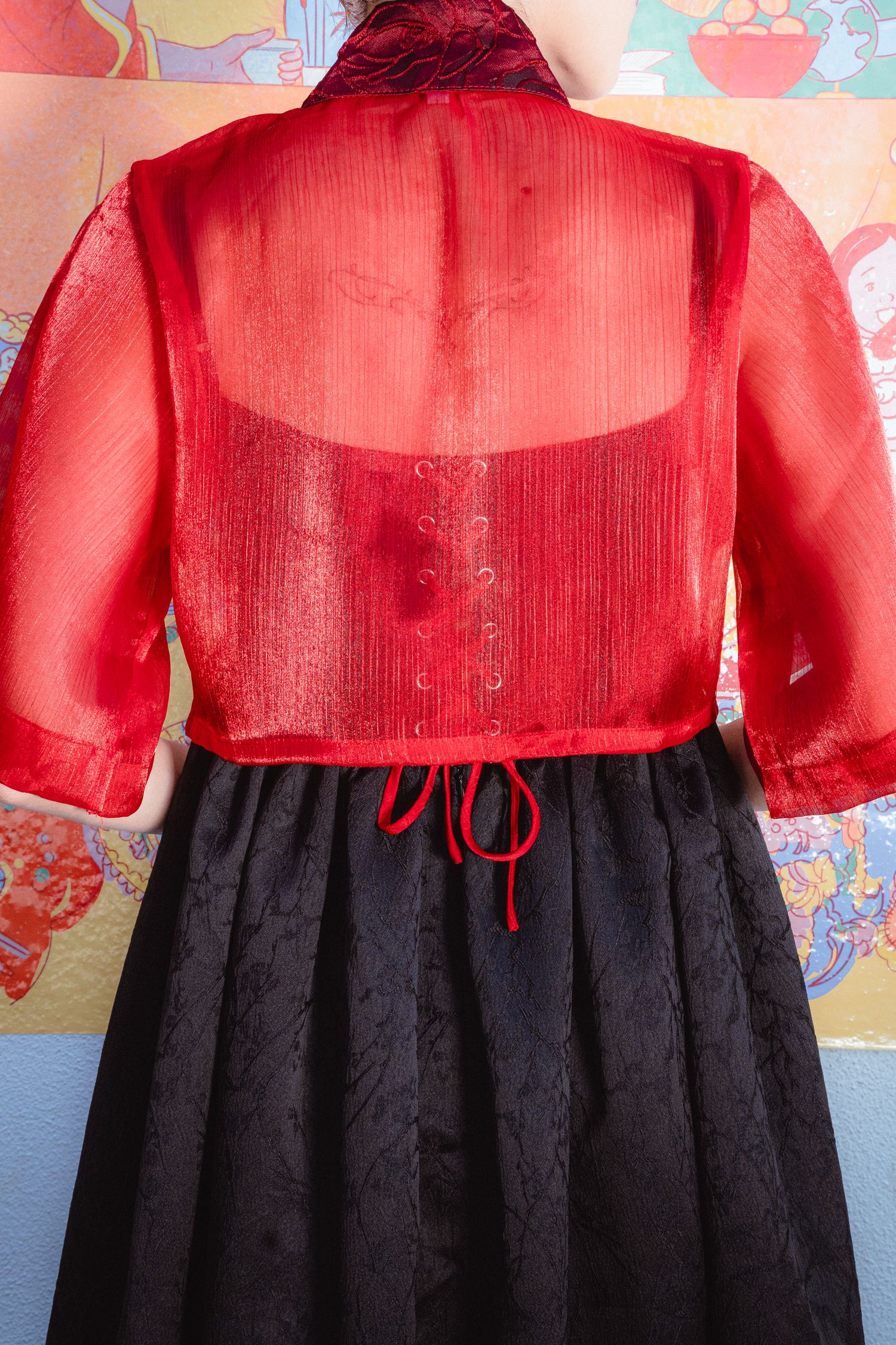 Red sheer organza bolero jacket with black brocade skirt, back view showing button closure and bow tie detail.