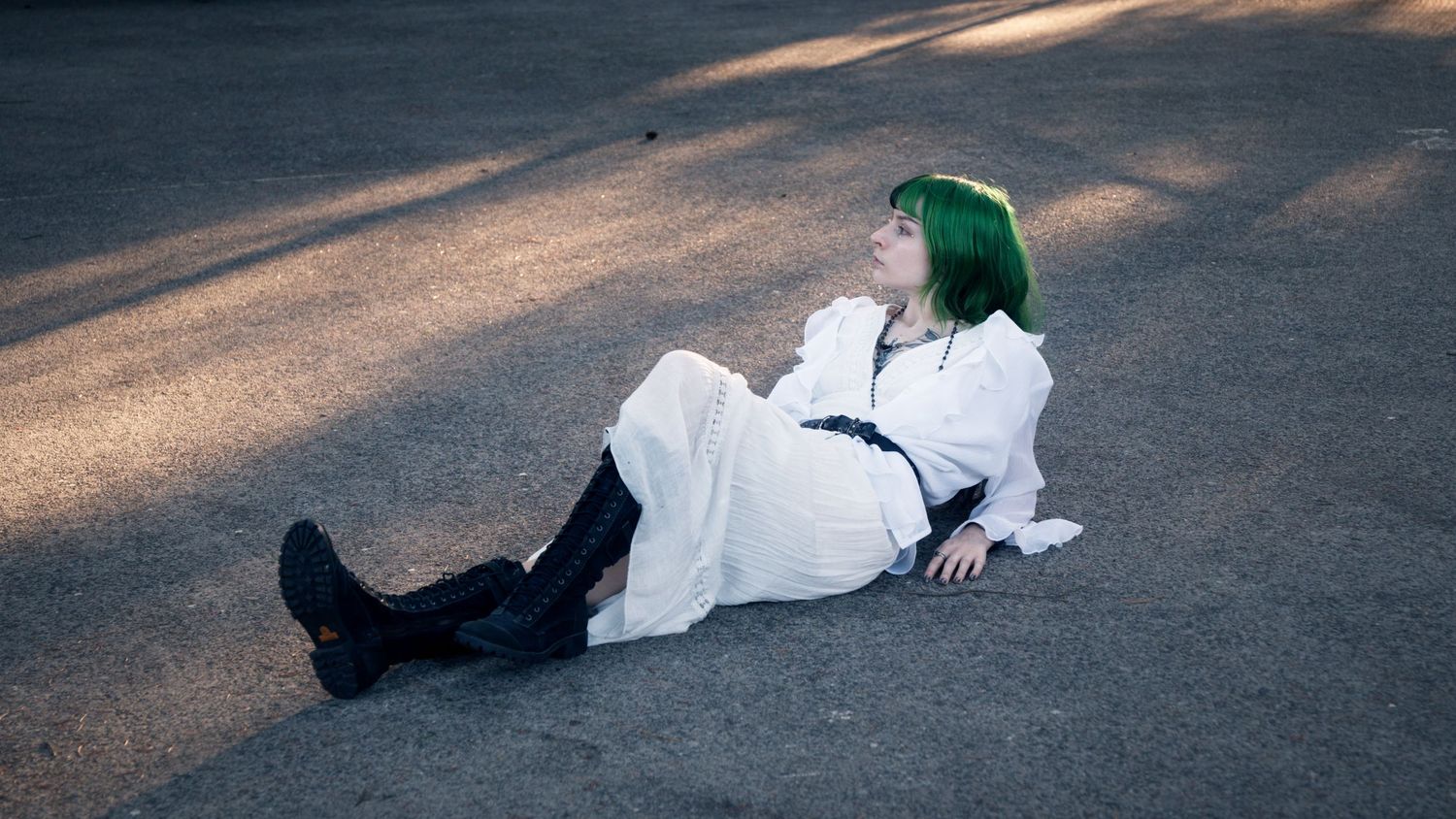 Woman with green hair in white dress and black boots reclines on sunlit pavement outdoors.