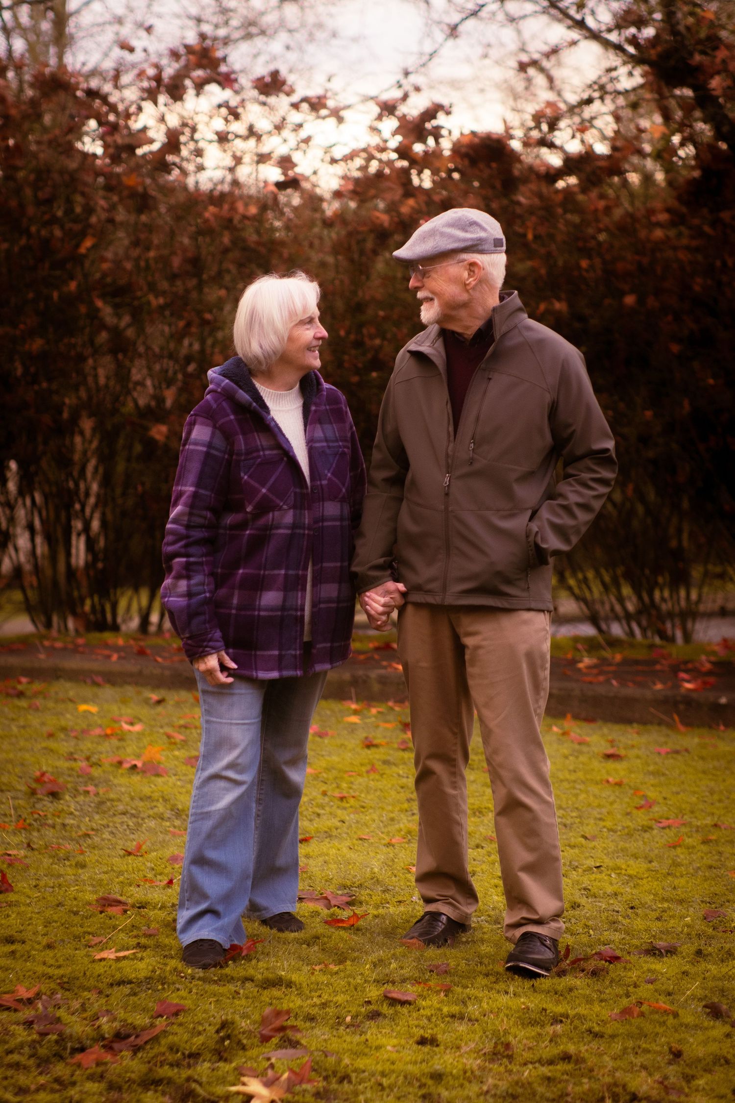 Elderly couple walking hand in hand on a grass lawn covered with fallen autumn leaves.
