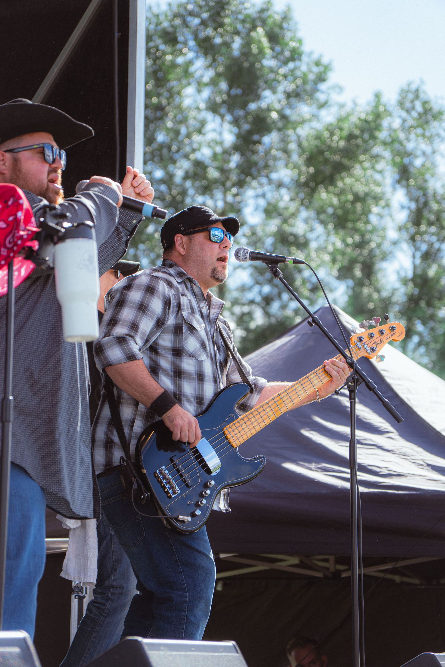 Bass guitarist in plaid shirt performs on outdoor stage alongside singer in black cowboy hat.
