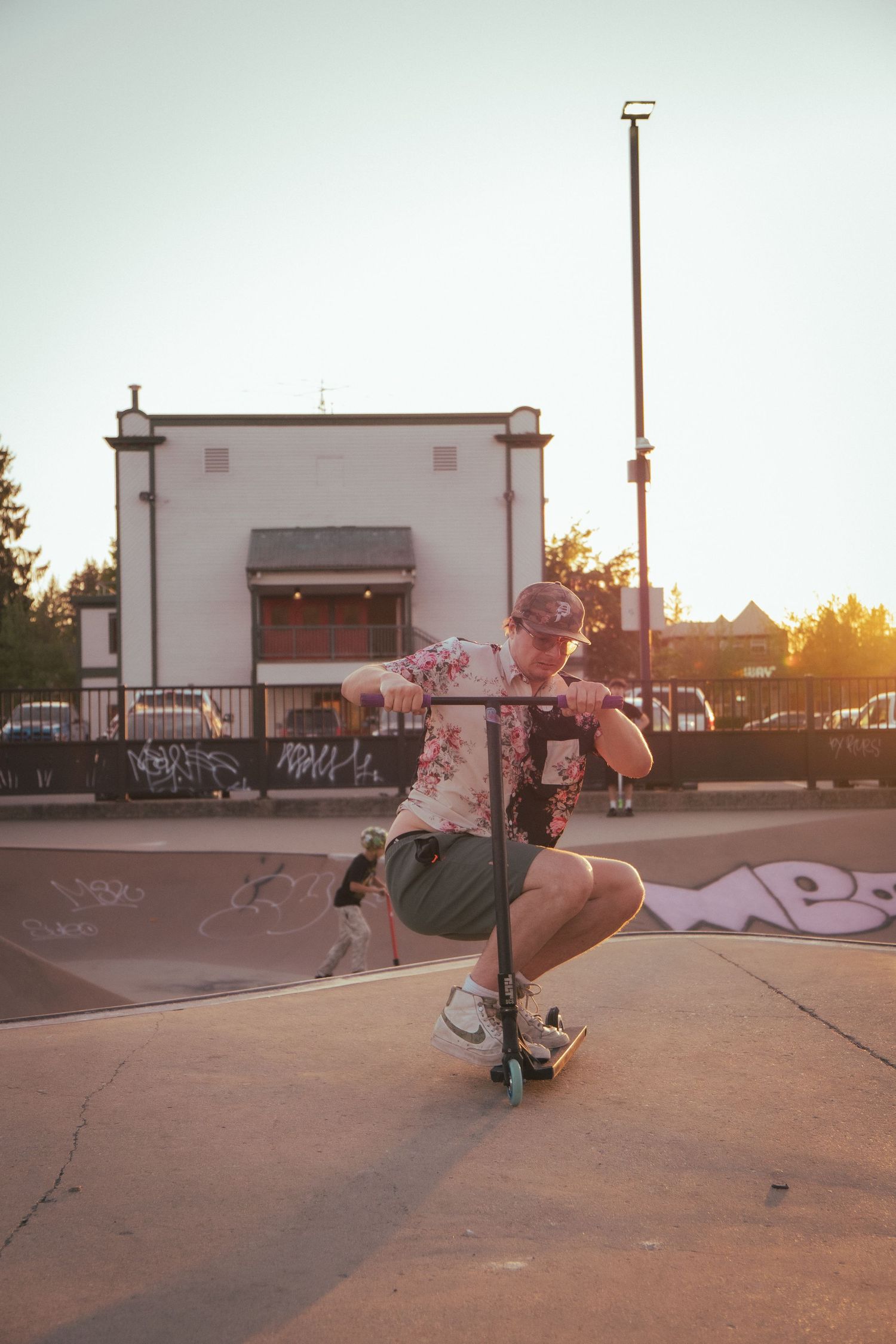 Young man crouching on a stunt scooter at an outdoor skate park during sunset.