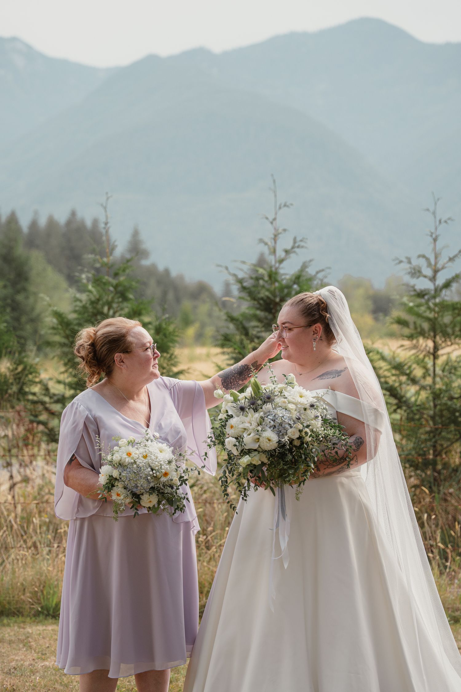 Bride in white gown and bridesmaid in lavender dress holding bouquets with mountain backdrop.