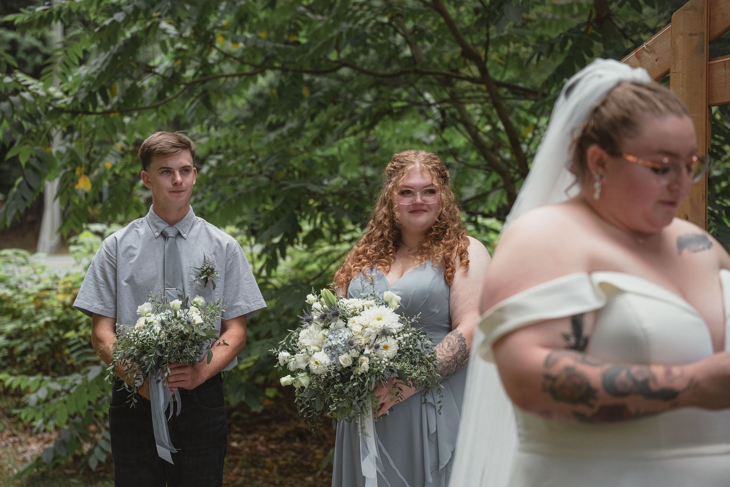 Bridesmaids in gray dresses holding white floral bouquets stand beside bride in white gown and veil outdoors.