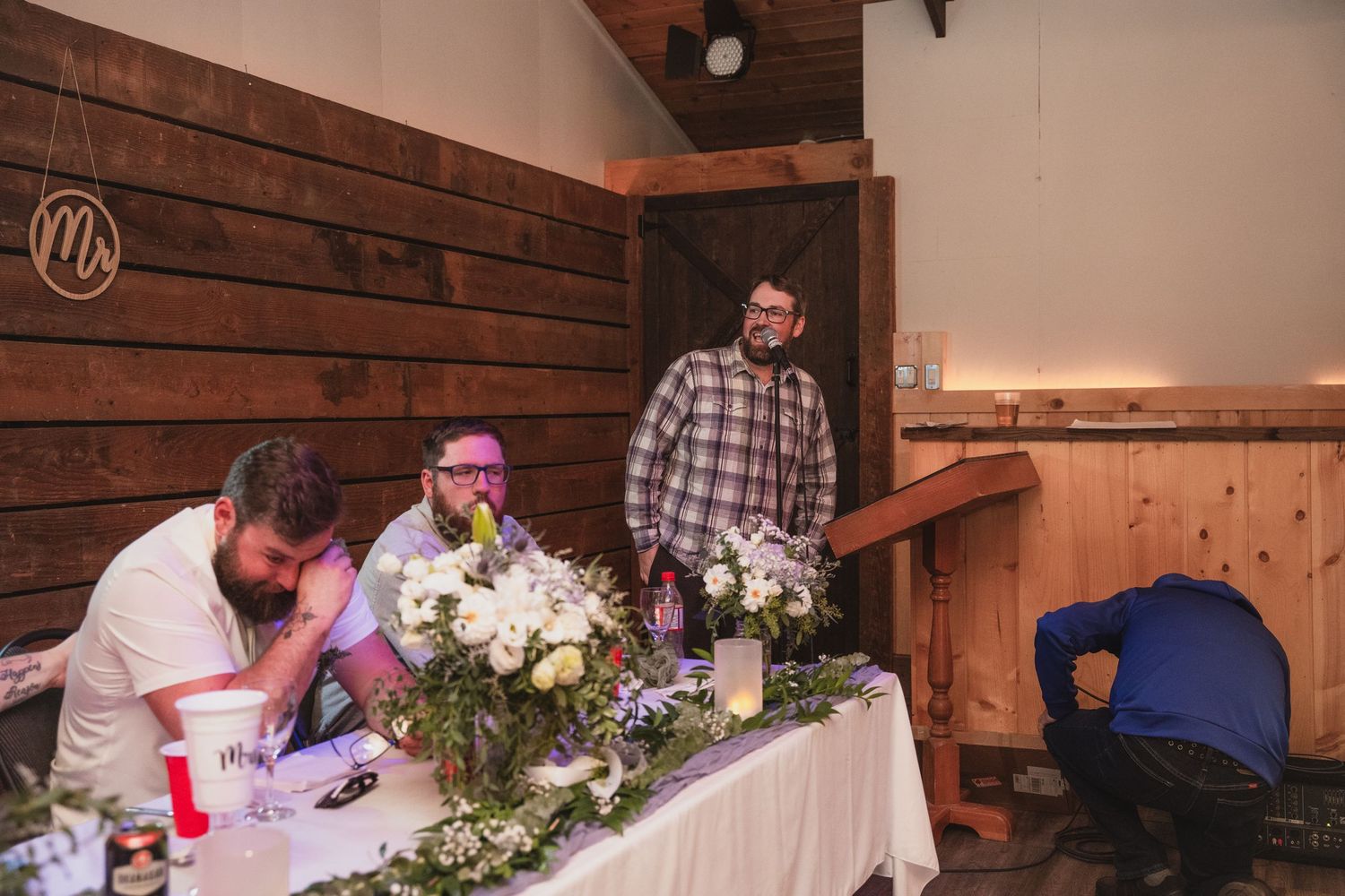 Groomsmen gather at a floral-decorated head table inside a rustic barn wedding venue.