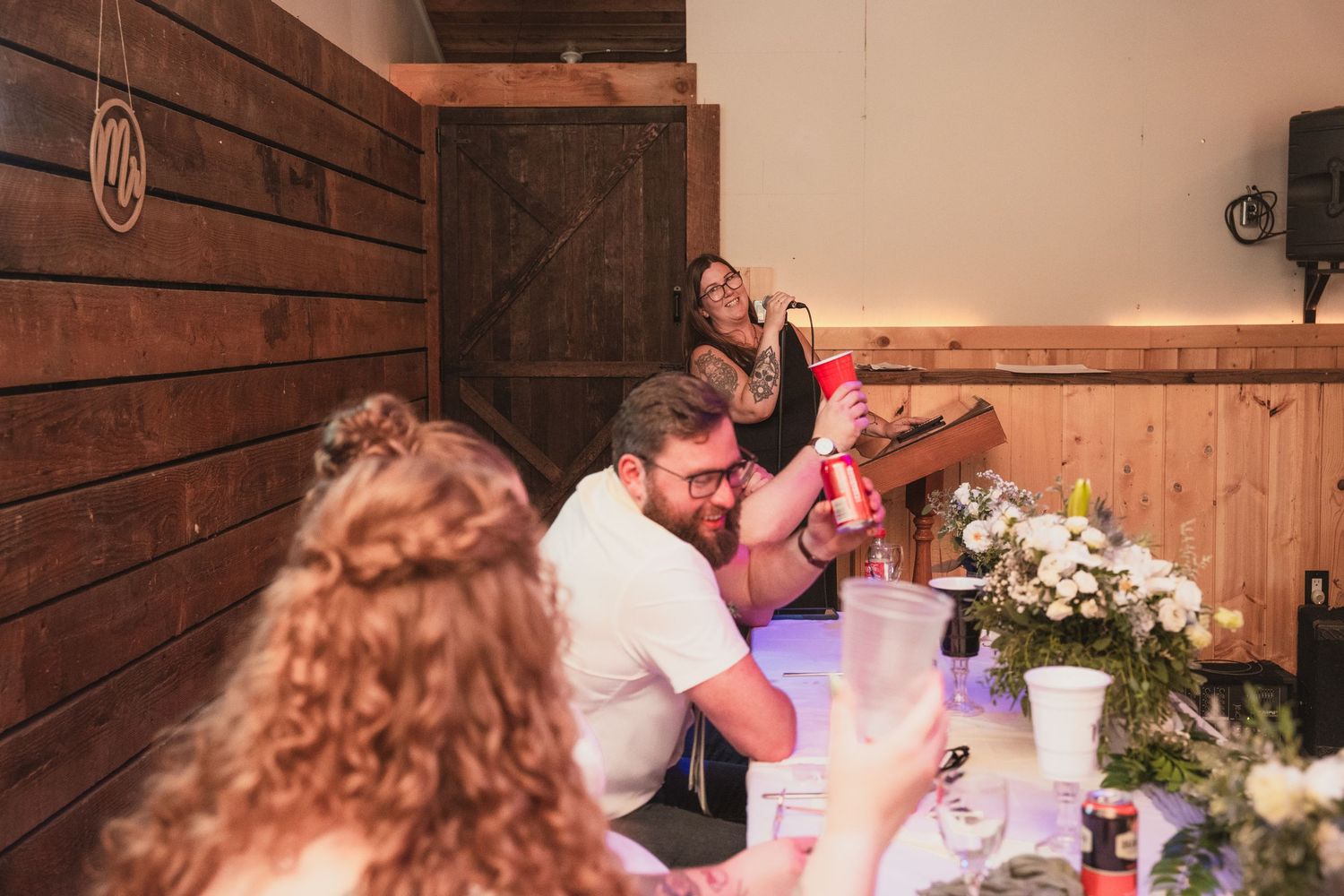 Group of people gathered around table with flowers and drinks in rustic barn setting.