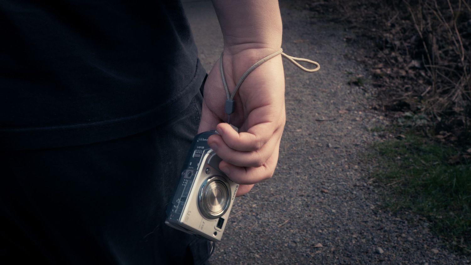 Close-up of a hand holding a compact silver digital camera with a wrist strap on a gravel path.