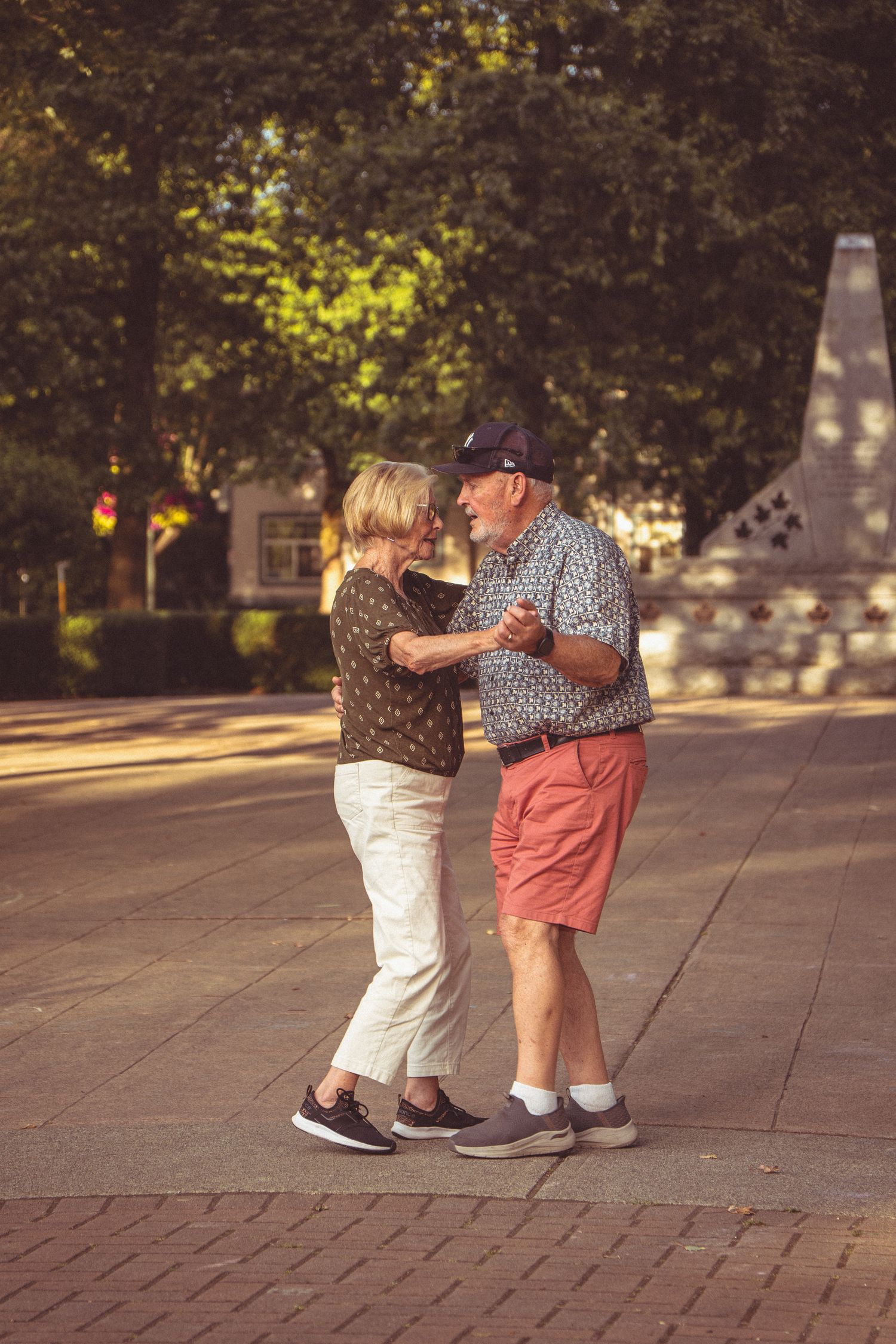 Elderly couple dancing together outdoors on a brick plaza surrounded by trees on a sunny day.