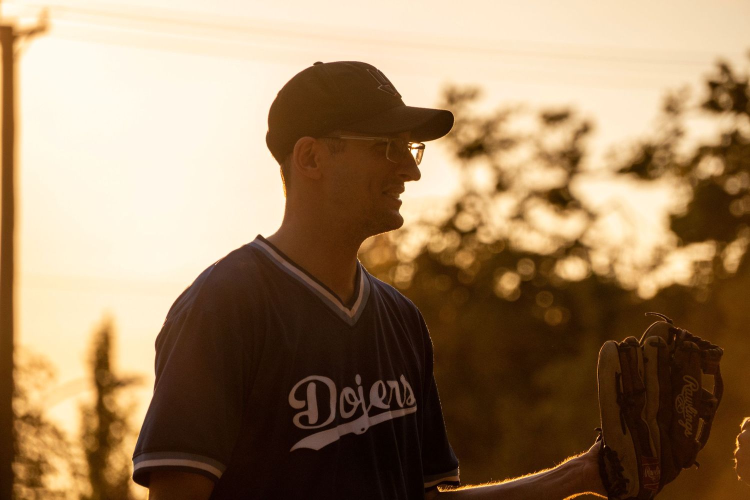 Dodgers softball player holding a glove stands in warm golden sunset light during an outdoor game.