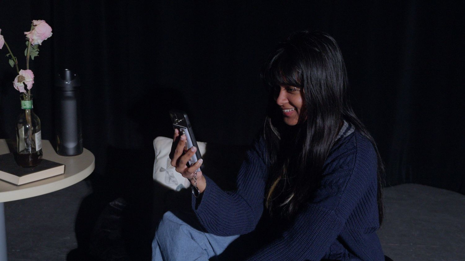 A woman with long dark hair sits on the floor looking at her phone in a dark setting.