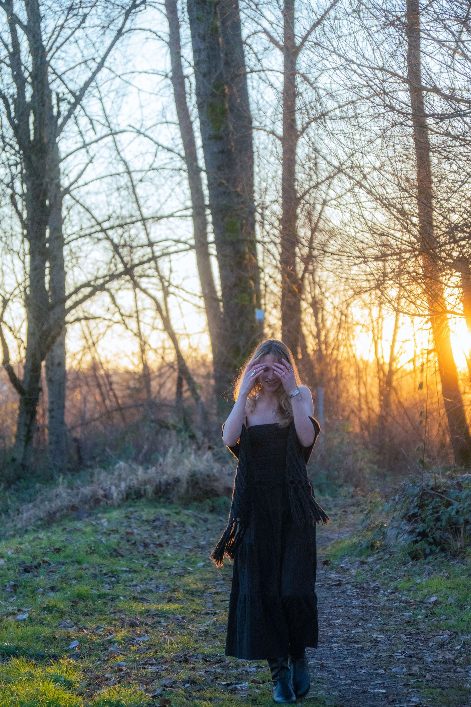 Blonde woman in black dress covering her face while standing on a forest path at sunset.
