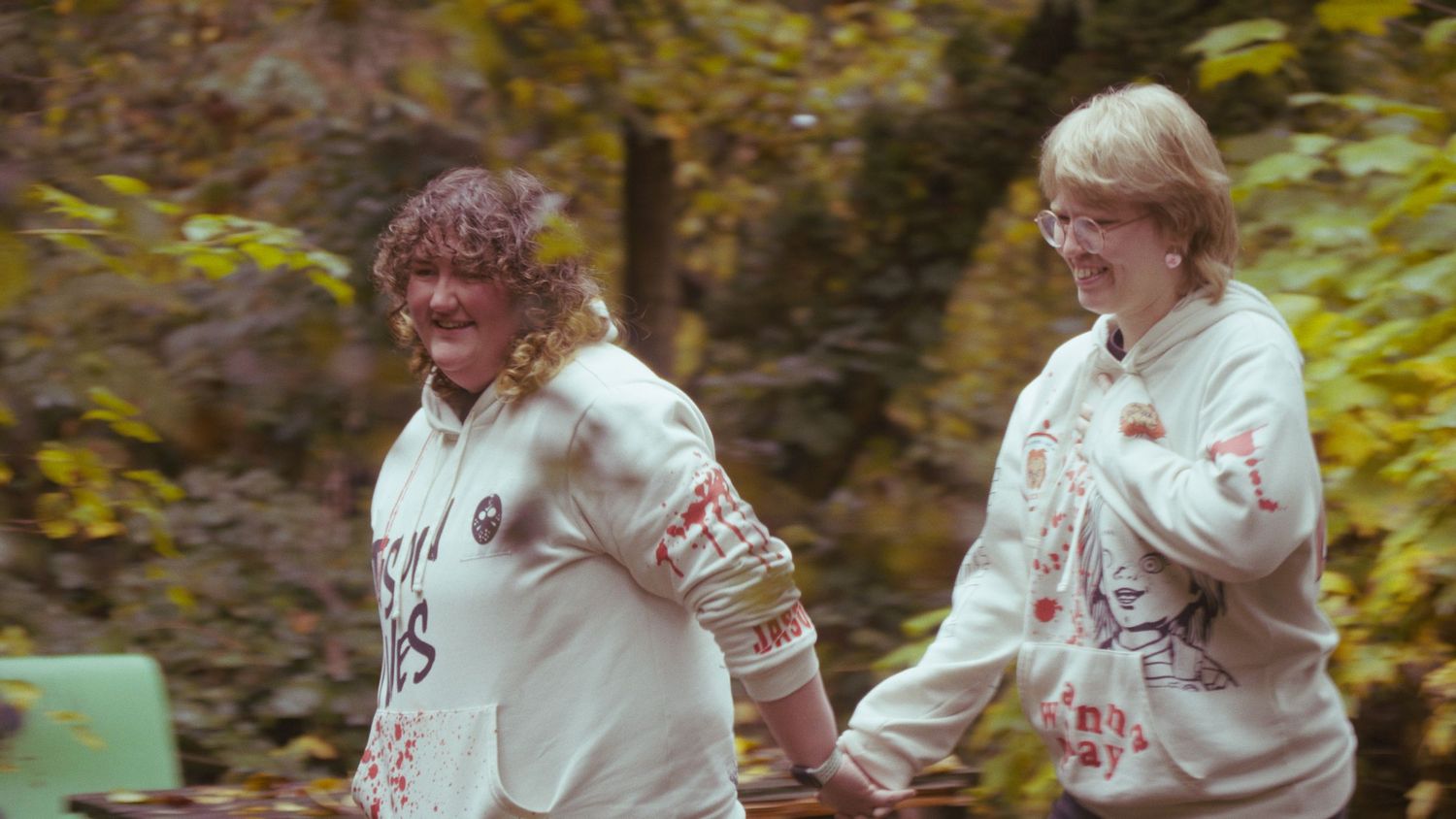 Two people in matching white hoodies walk together through a colorful autumn forest setting.