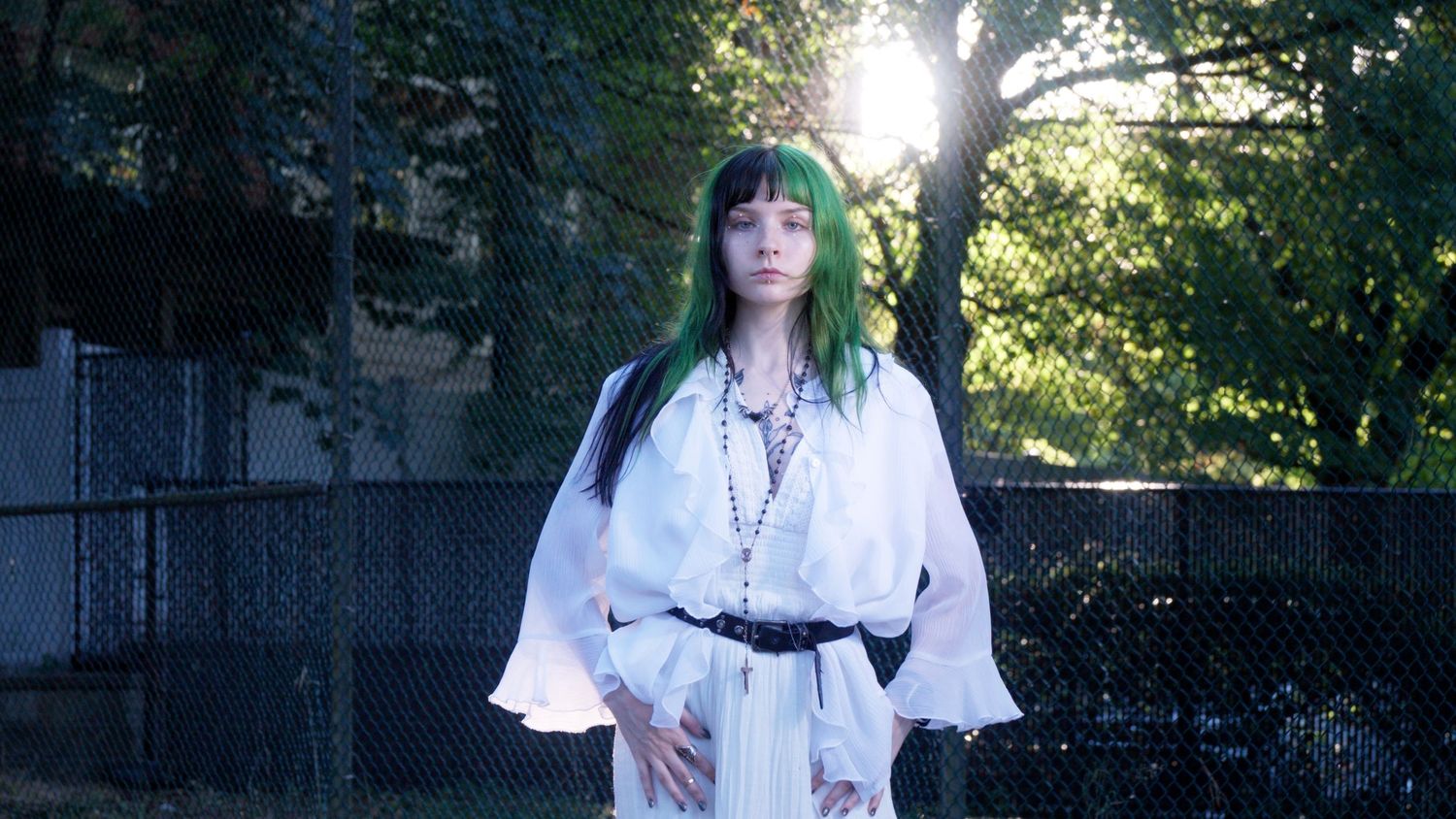 Woman with green hair in white robe and black belt stands outdoors near fence in sunlight.