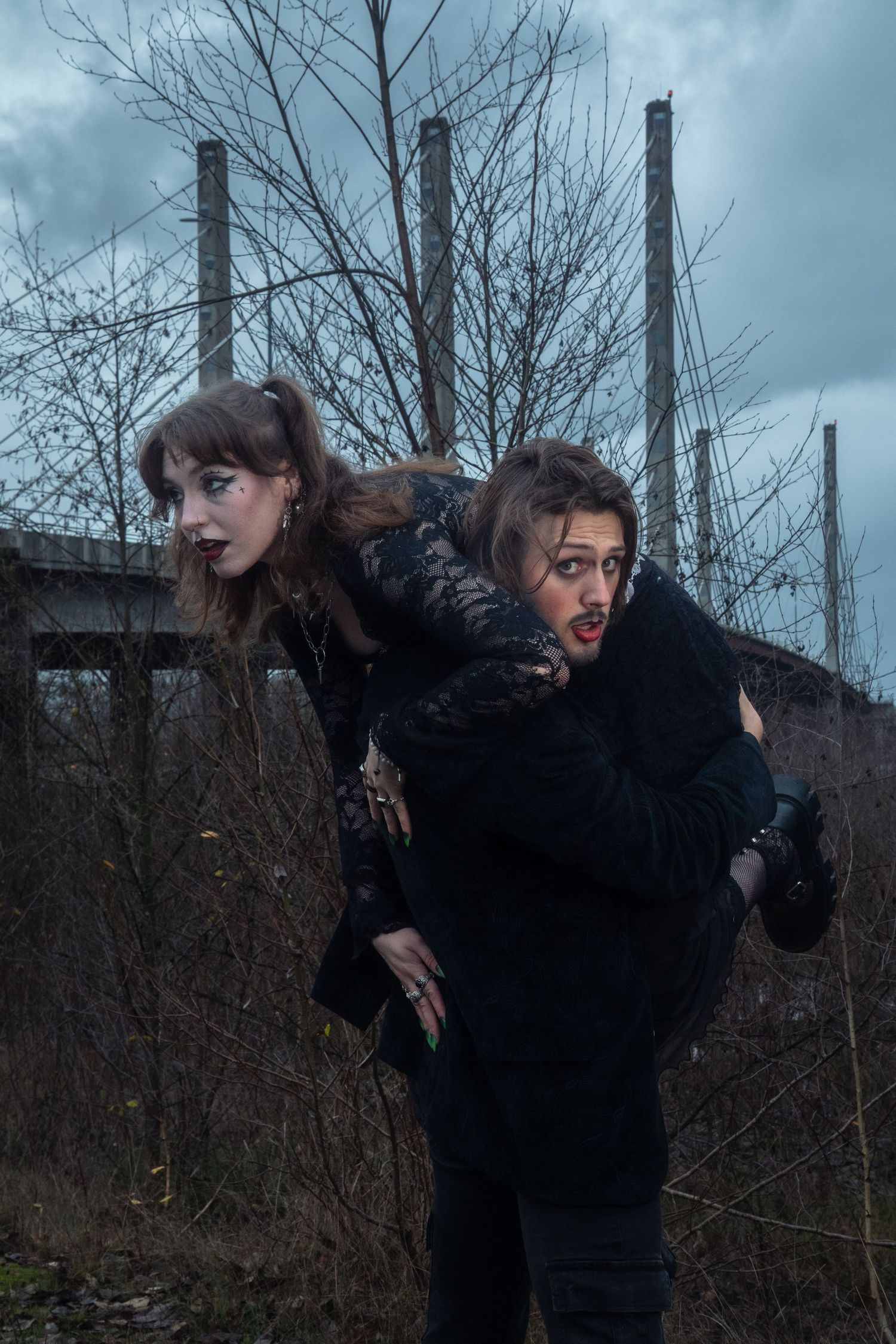 Gothic couple in black playfully pose with piggyback ride among bare trees on overcast day.