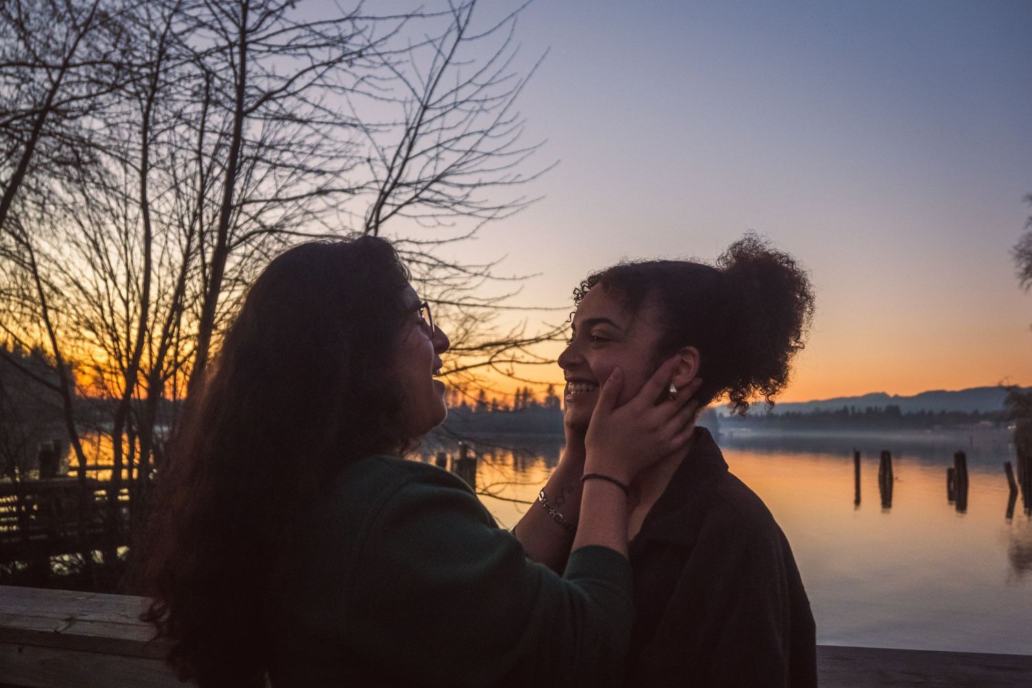 Two women share a tender romantic moment by a lakeside at golden sunset, one gently cradling the others face.