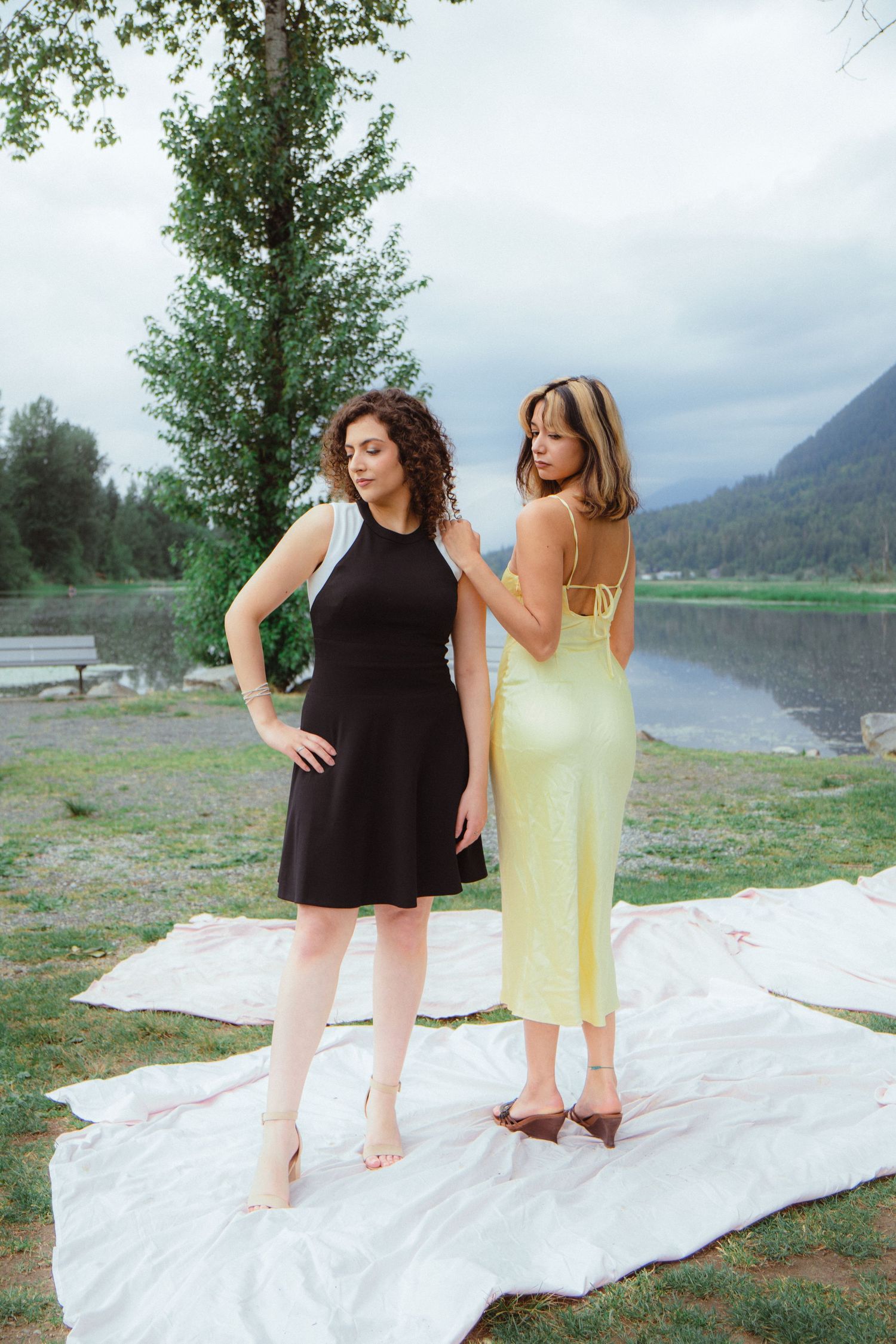 Two women in elegant dresses pose on a white blanket by a mountain lake, one in black and one in yellow.