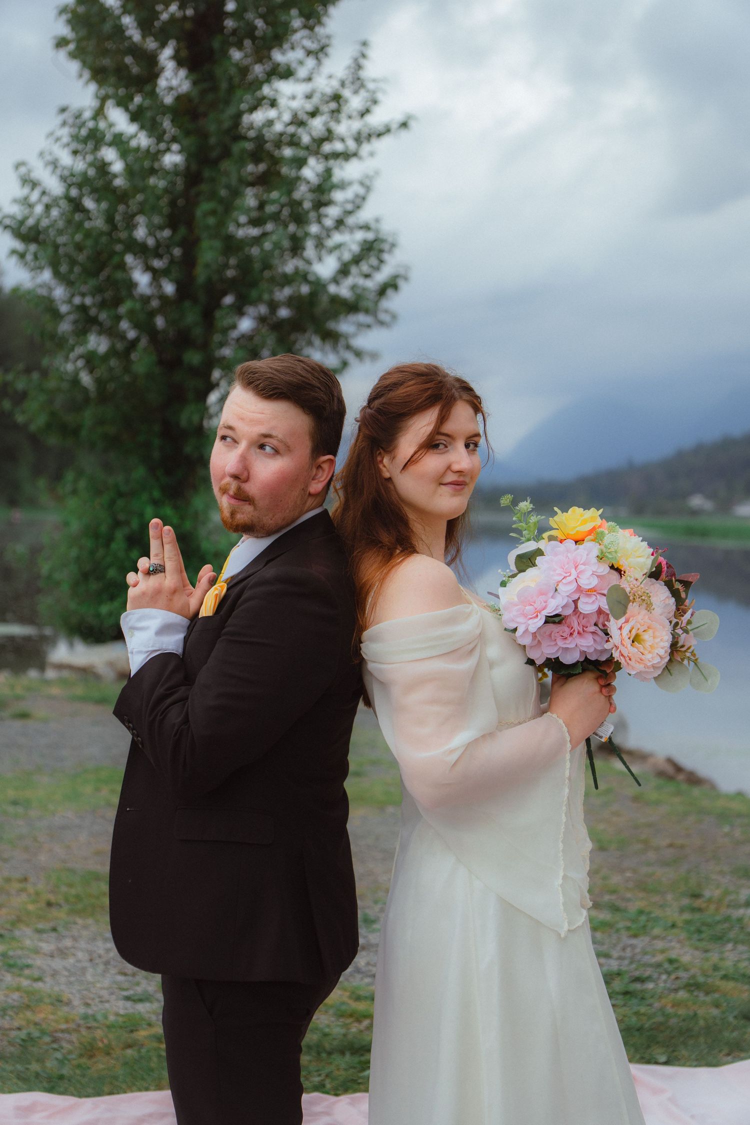 Bride and groom pose back-to-back with playful expressions by a mountain lake.