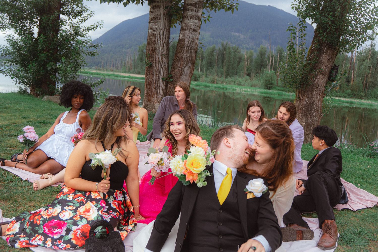 Bride and groom share a romantic kiss surrounded by wedding guests on a lakeside blanket.