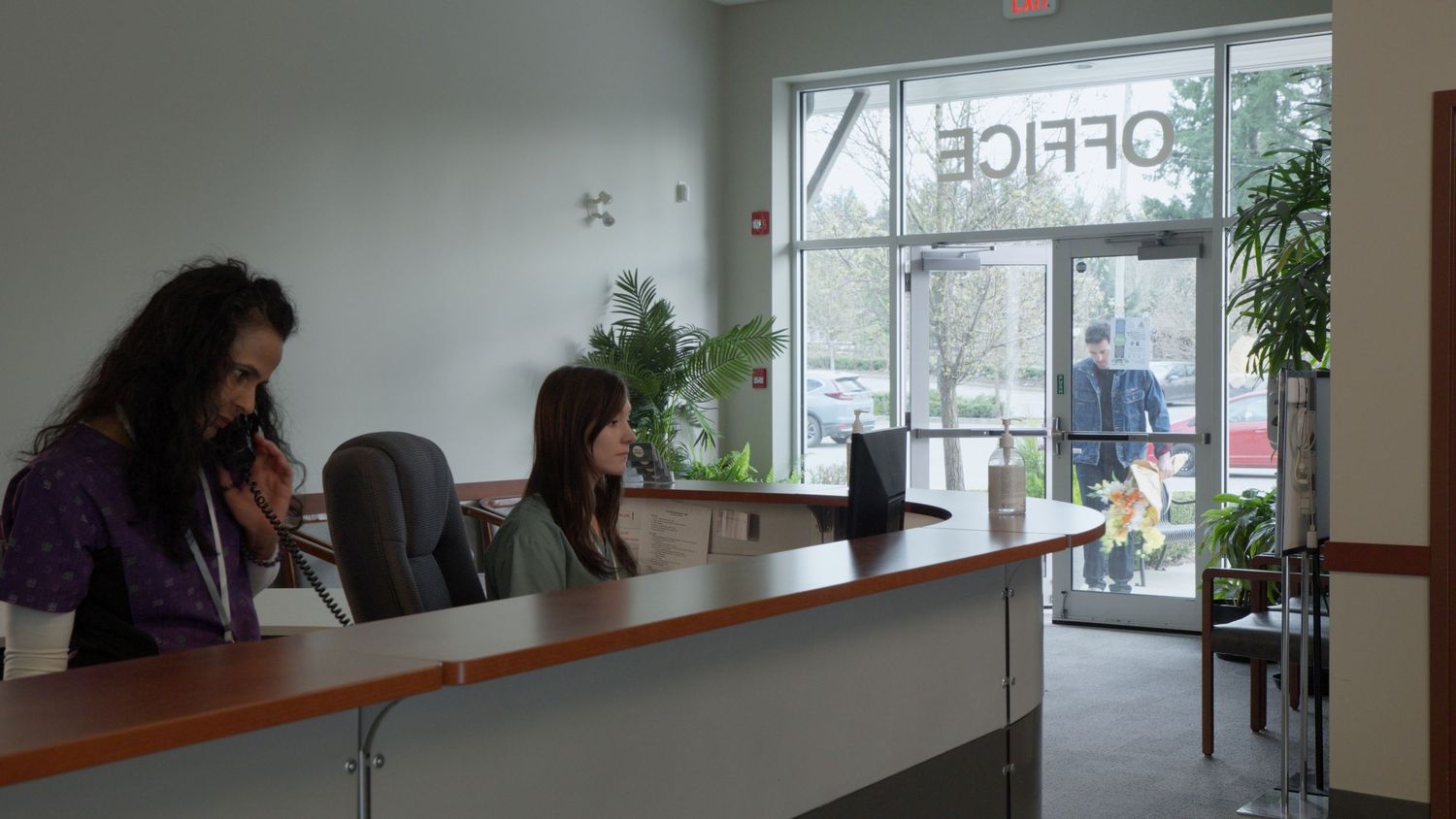 Two women sit at an office reception desk with plants and large windows in a bright modern office.