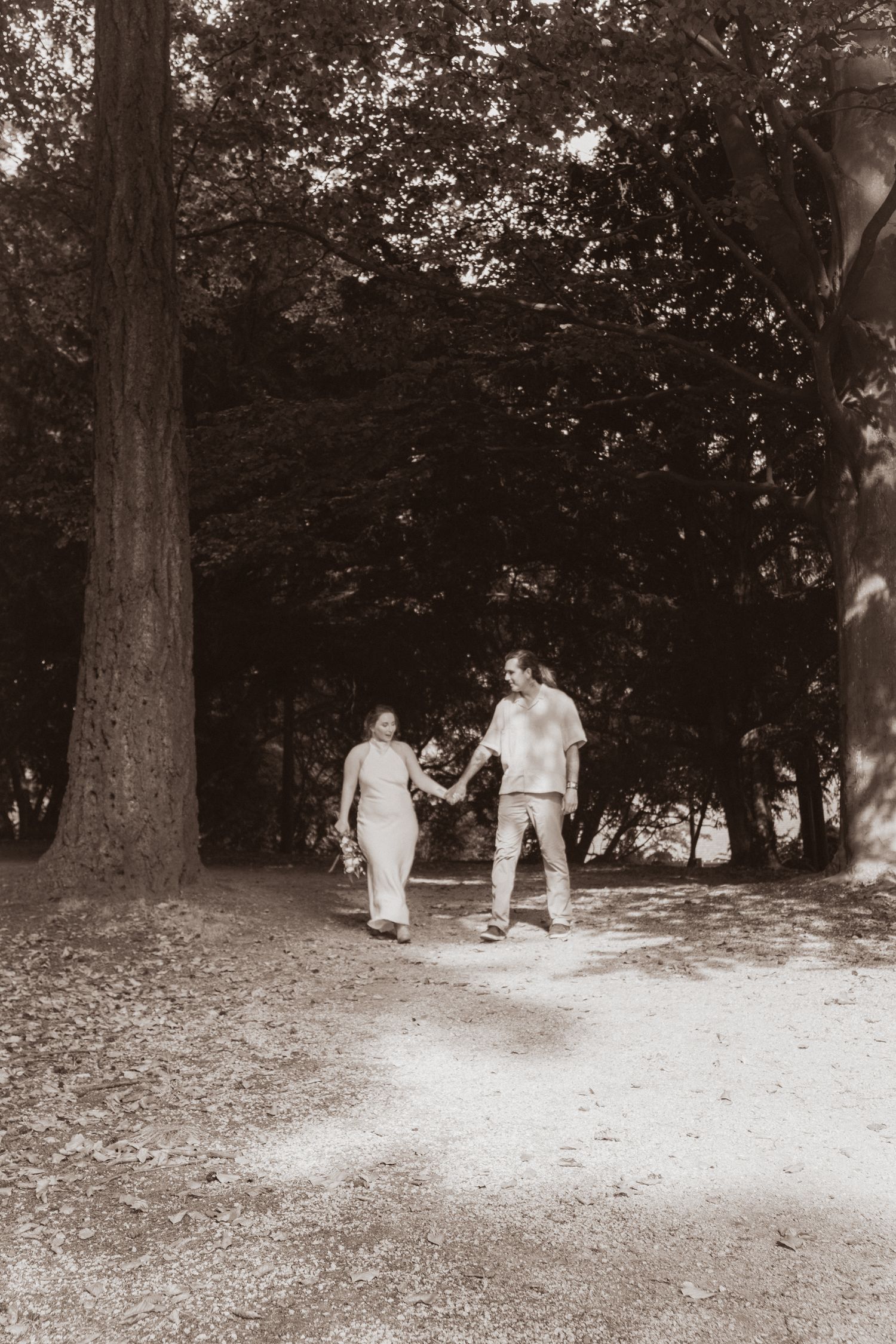 Sepia-toned image of bride and groom holding hands while walking on a sunlit forest path.
