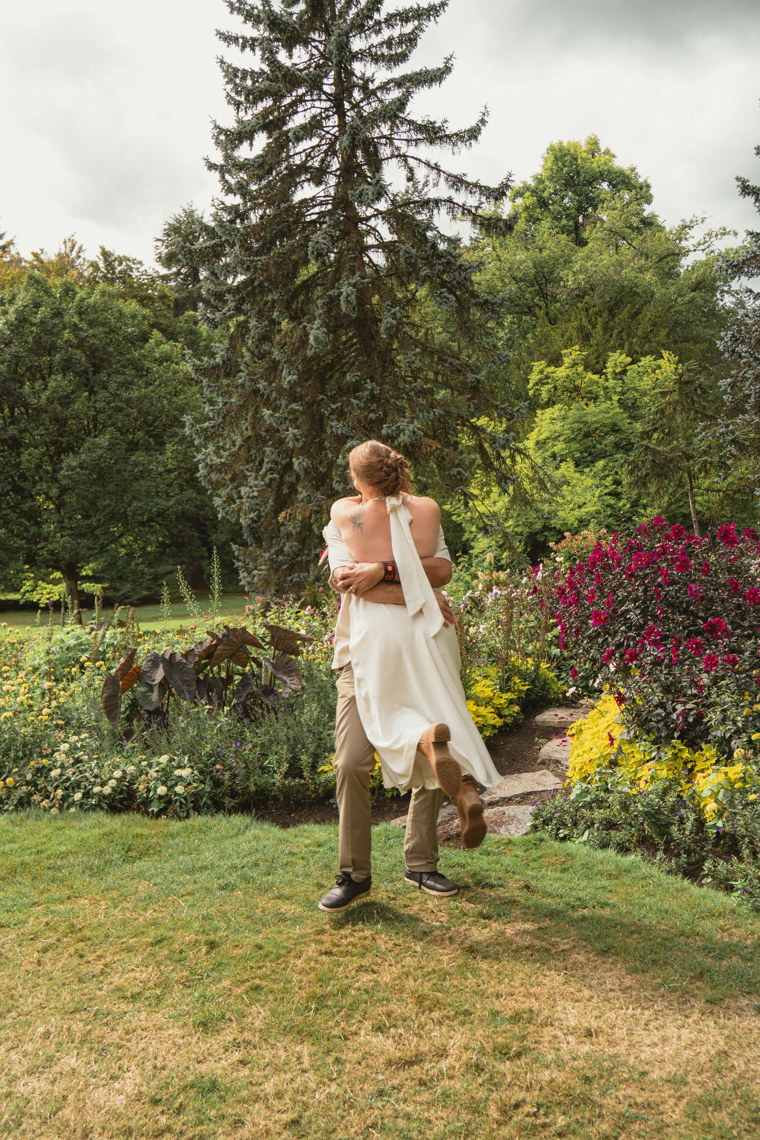 Groom lifting bride in white dress, spinning joyfully in a lush garden with colorful flowers and tall trees.