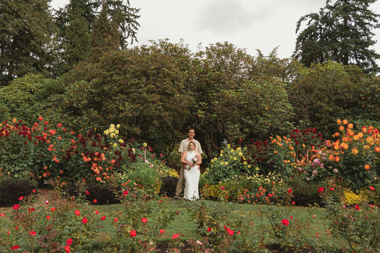 Couple poses in a lush garden wedding setting surrounded by vibrant red, orange, and yellow roses with tall green trees.