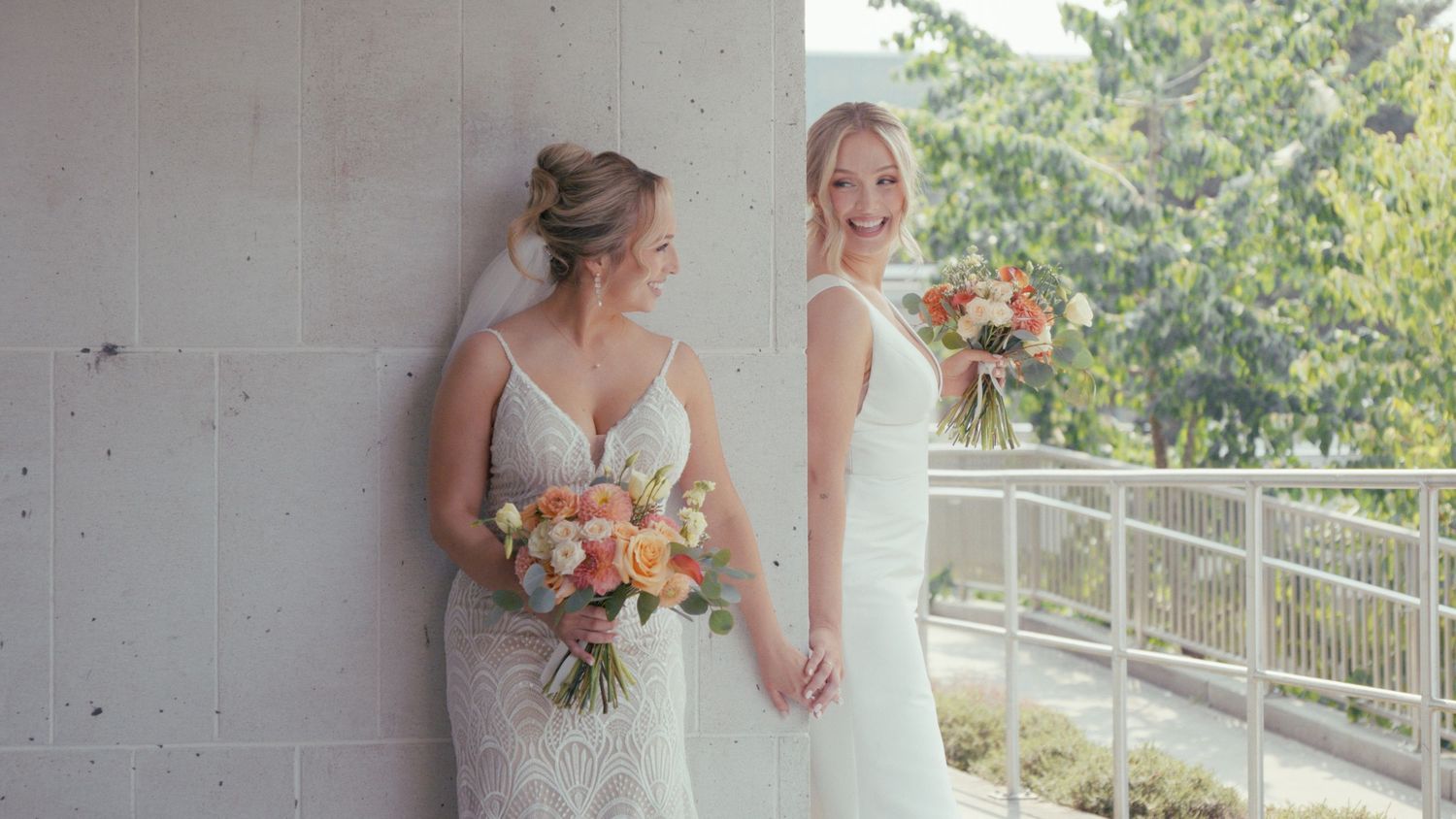 Two brides in white wedding gowns holding bouquets of peach and coral flowers on an outdoor balcony.