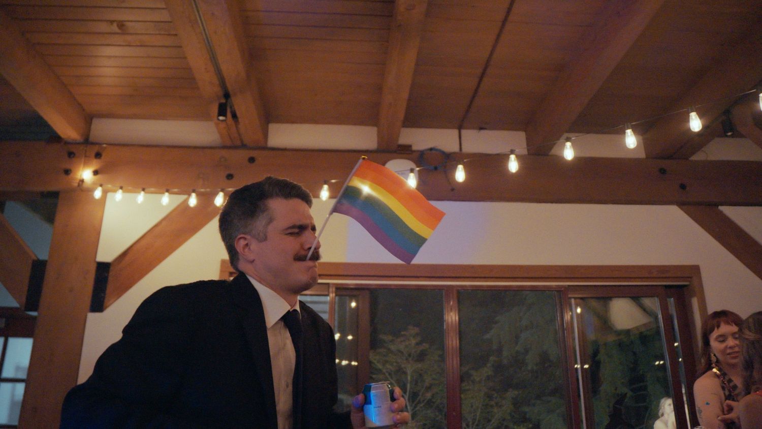 Man in suit waving a rainbow Pride flag at an indoor celebration with string lights and wooden beams.