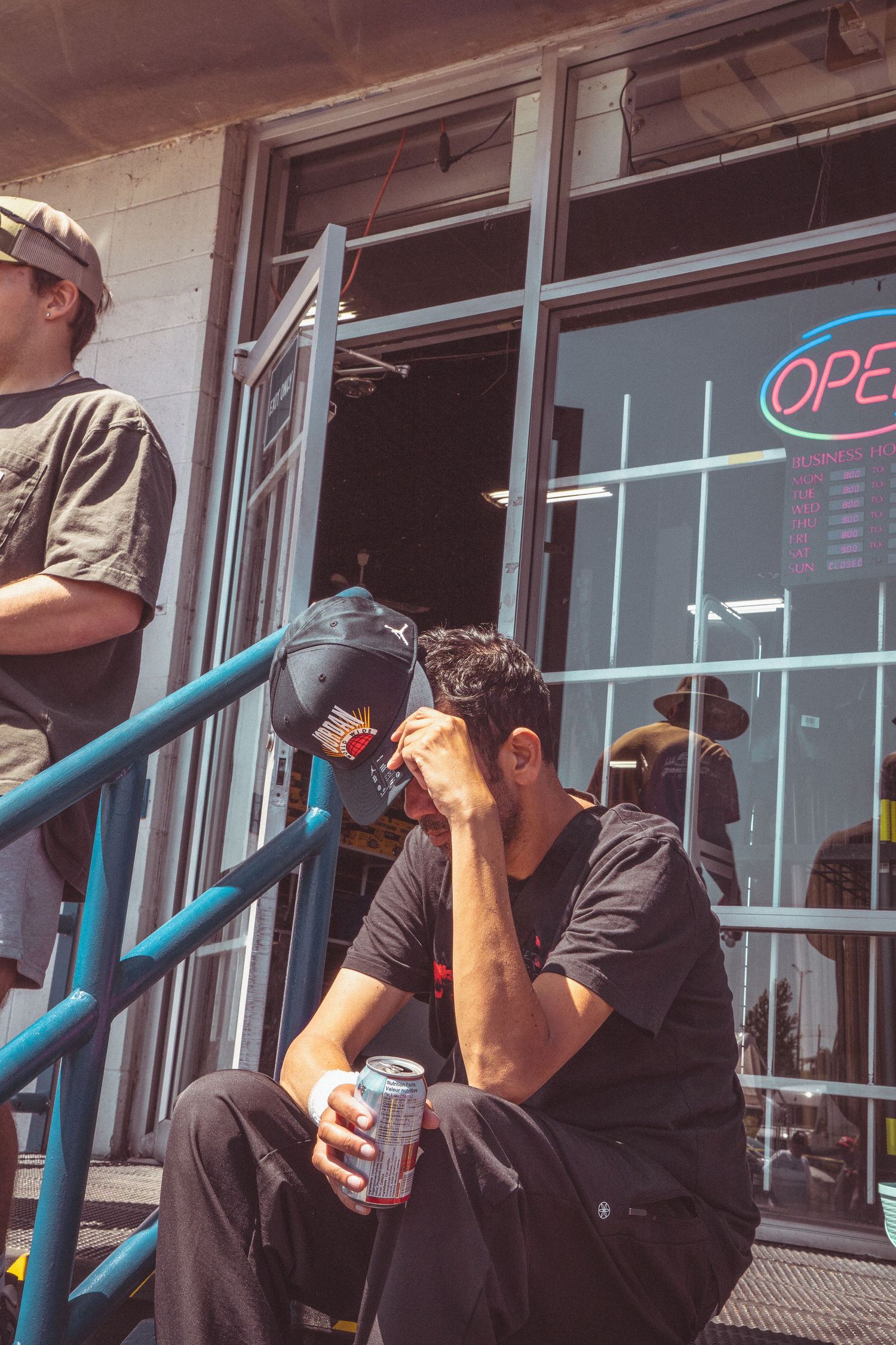 Man in black shirt sitting on steps holding a can, head in hand, outside an open shop.