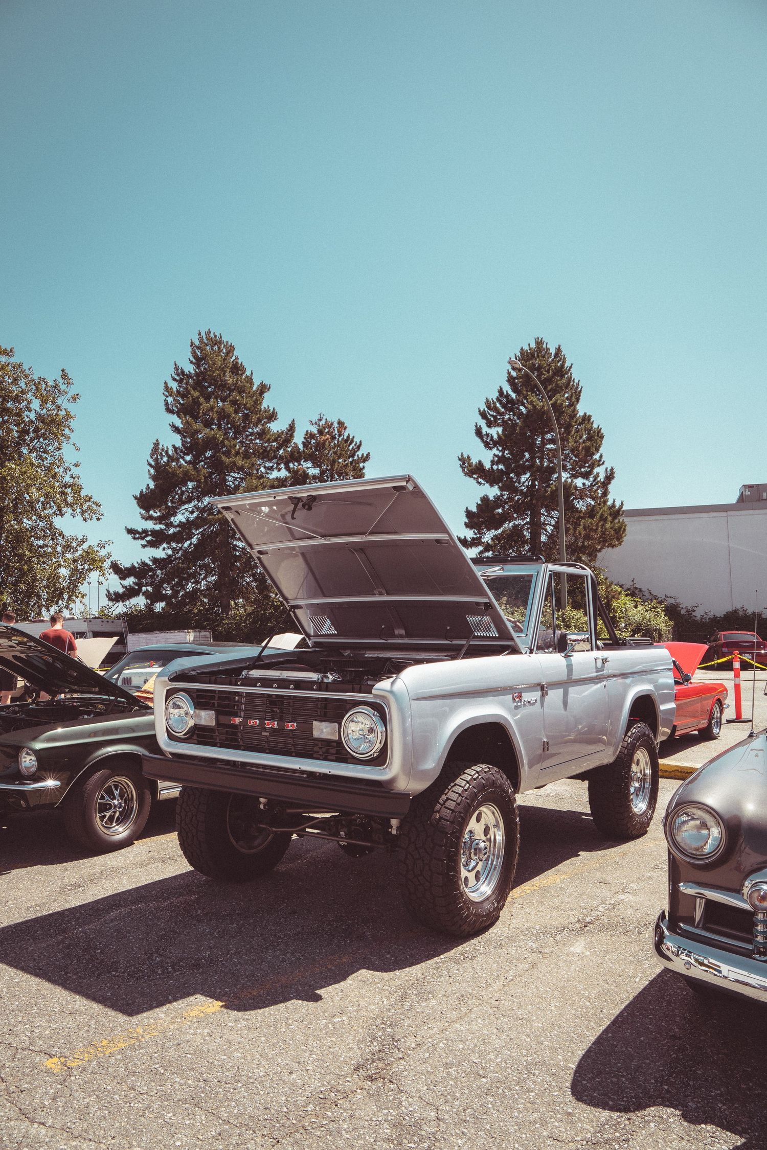 Silver vintage Ford Bronco with hood open displayed at an outdoor classic car show on a sunny day.
