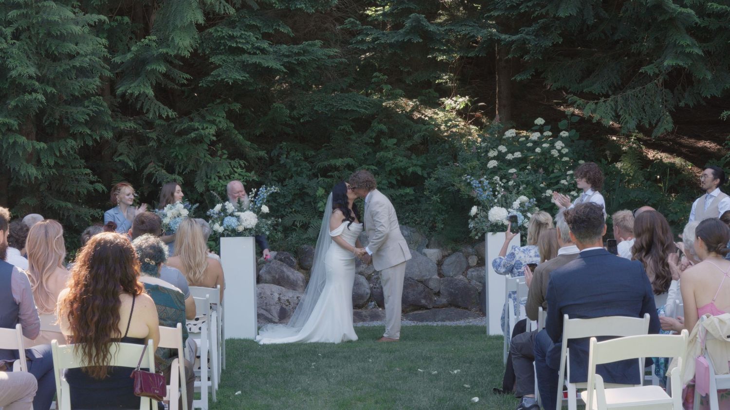 Bride and groom kiss at altar during outdoor wedding ceremony with seated guests watching.