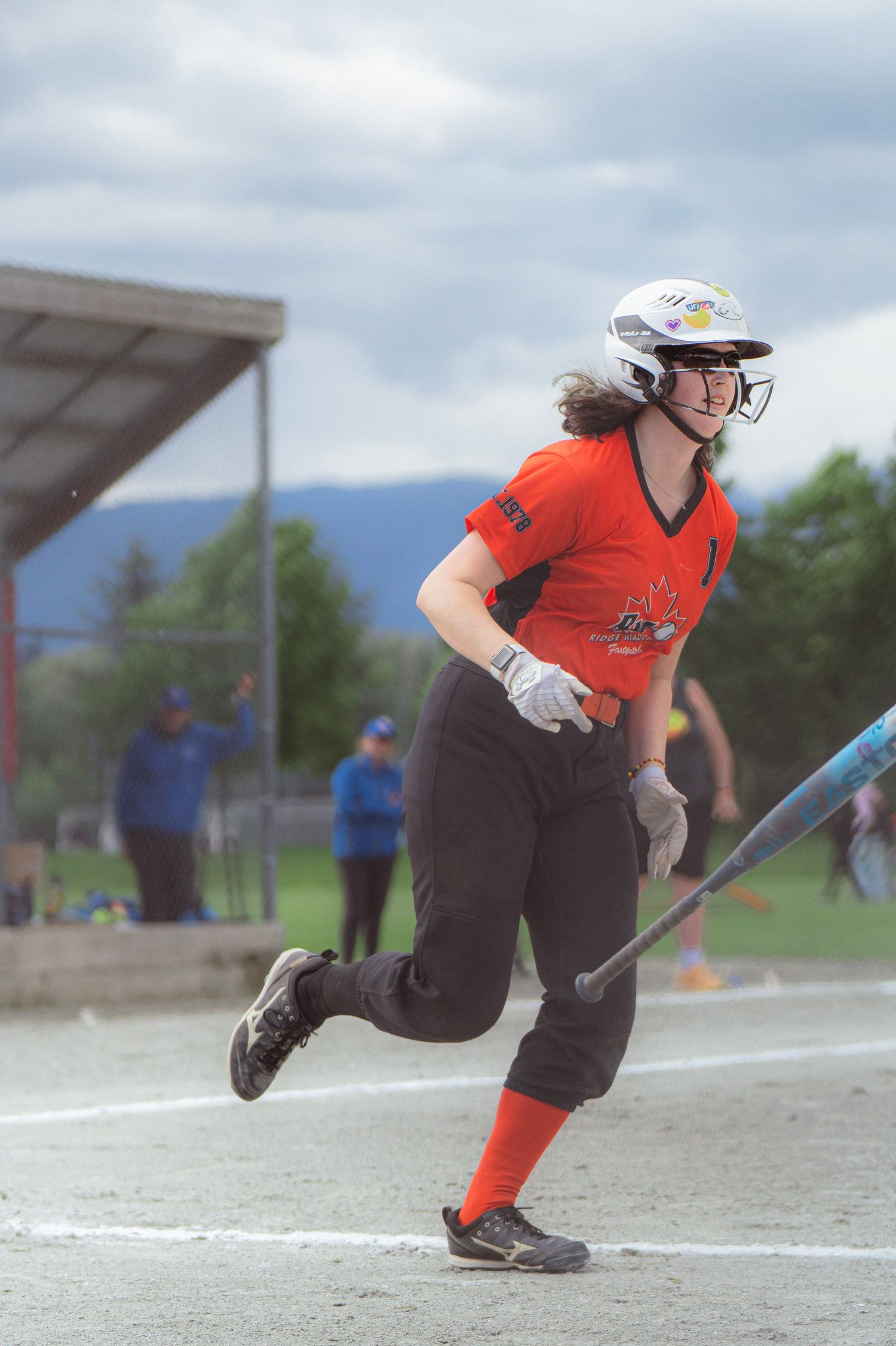 Softball player in orange uniform runs to first base after hitting the ball during a game.
