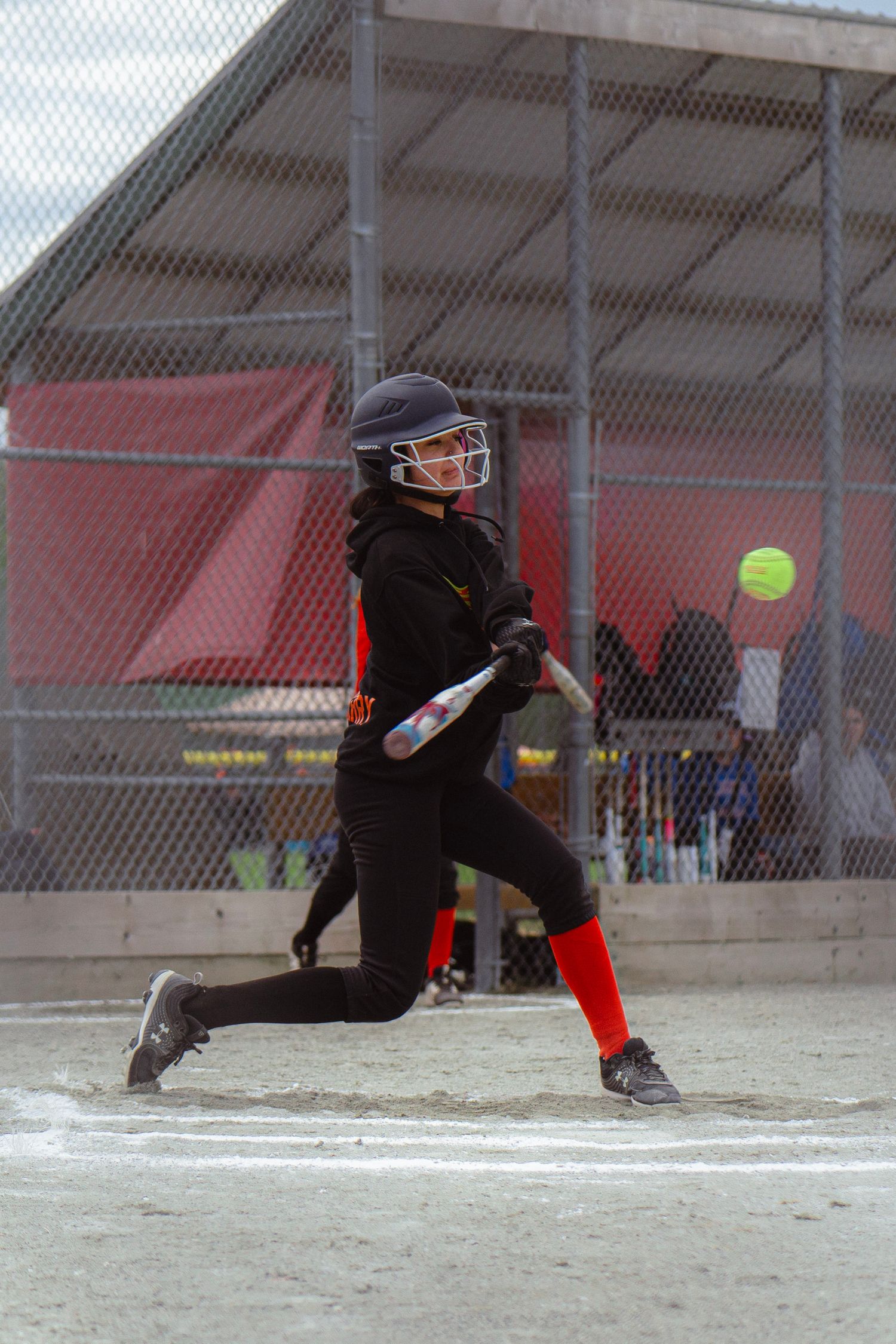 Softball player in black uniform swinging at a yellow ball near home plate.