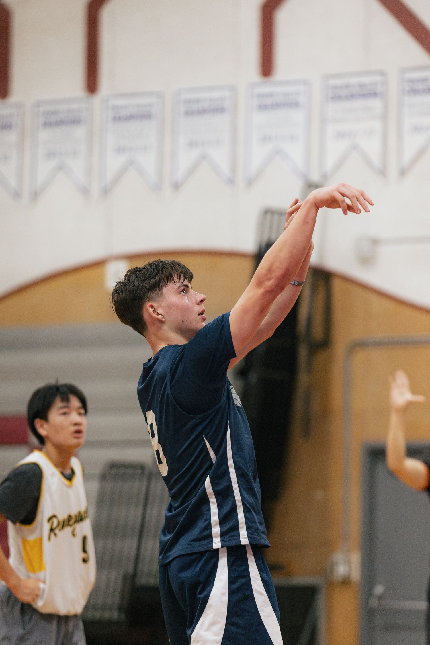 Young basketball player in navy uniform releases a jump shot during indoor game.