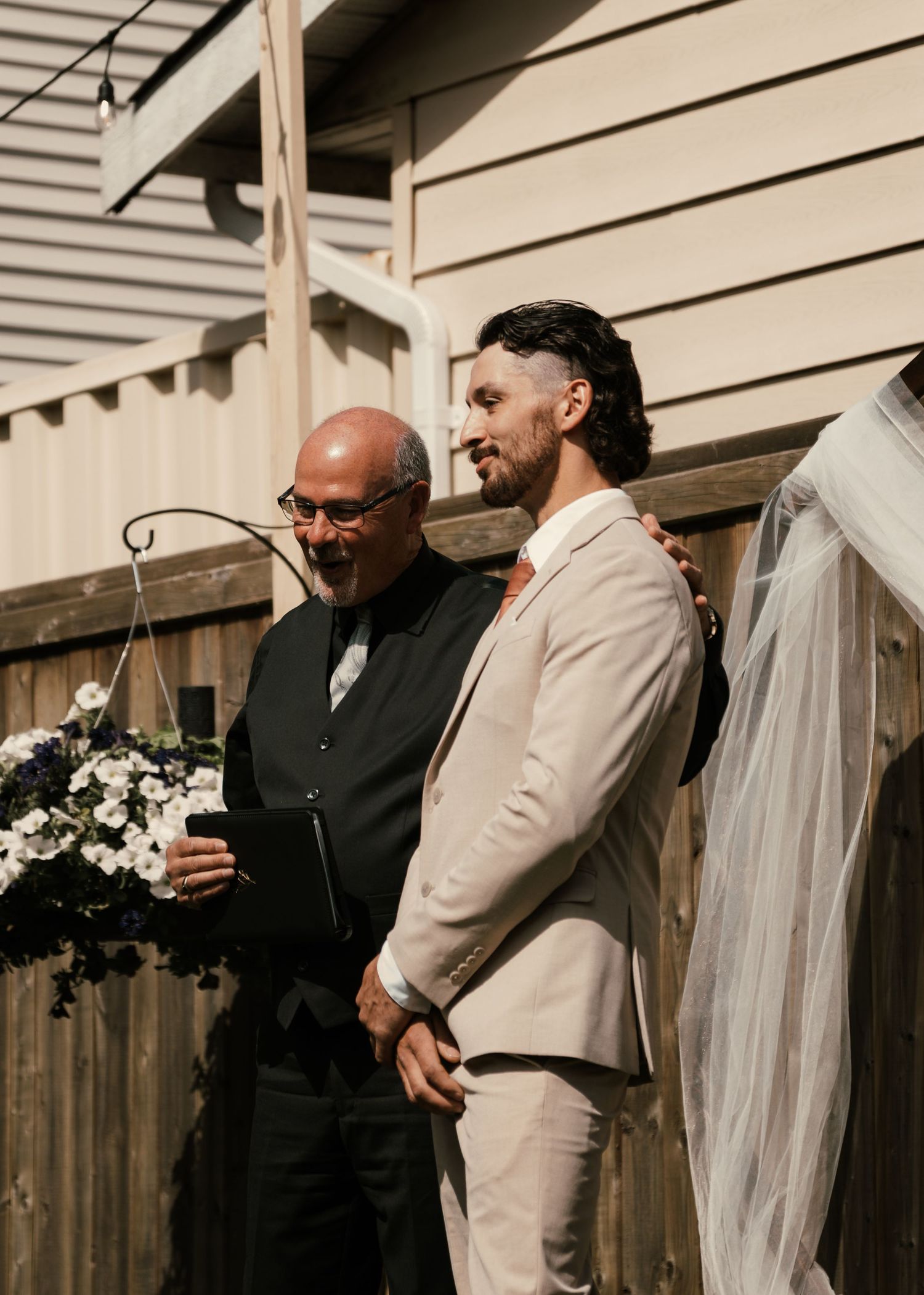 Groom in beige suit stands with officiant reading vows at outdoor wedding ceremony near wooden fence.
