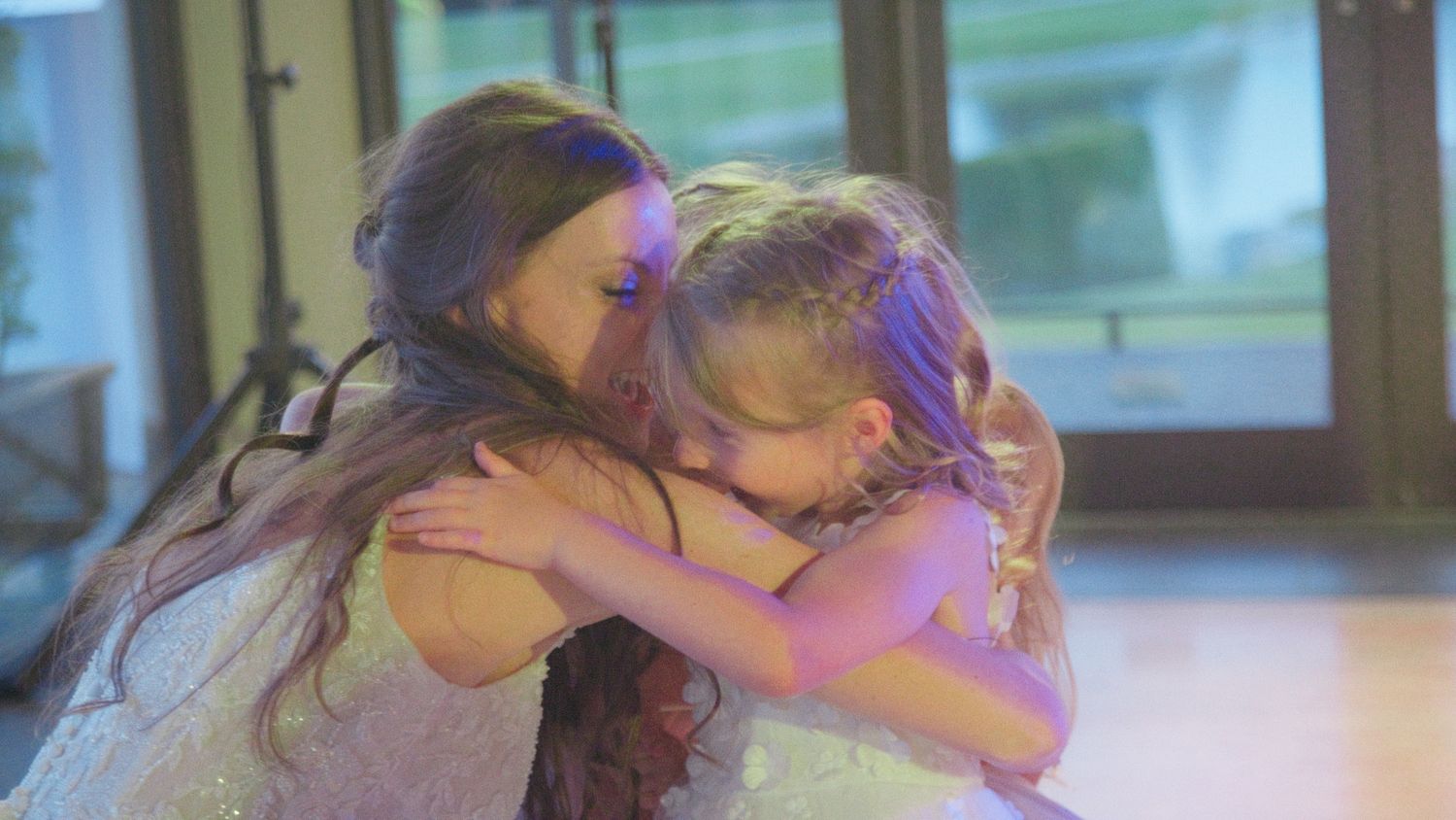 Bride shares a heartfelt hug with a young flower girl in a sparkly dress during wedding reception.