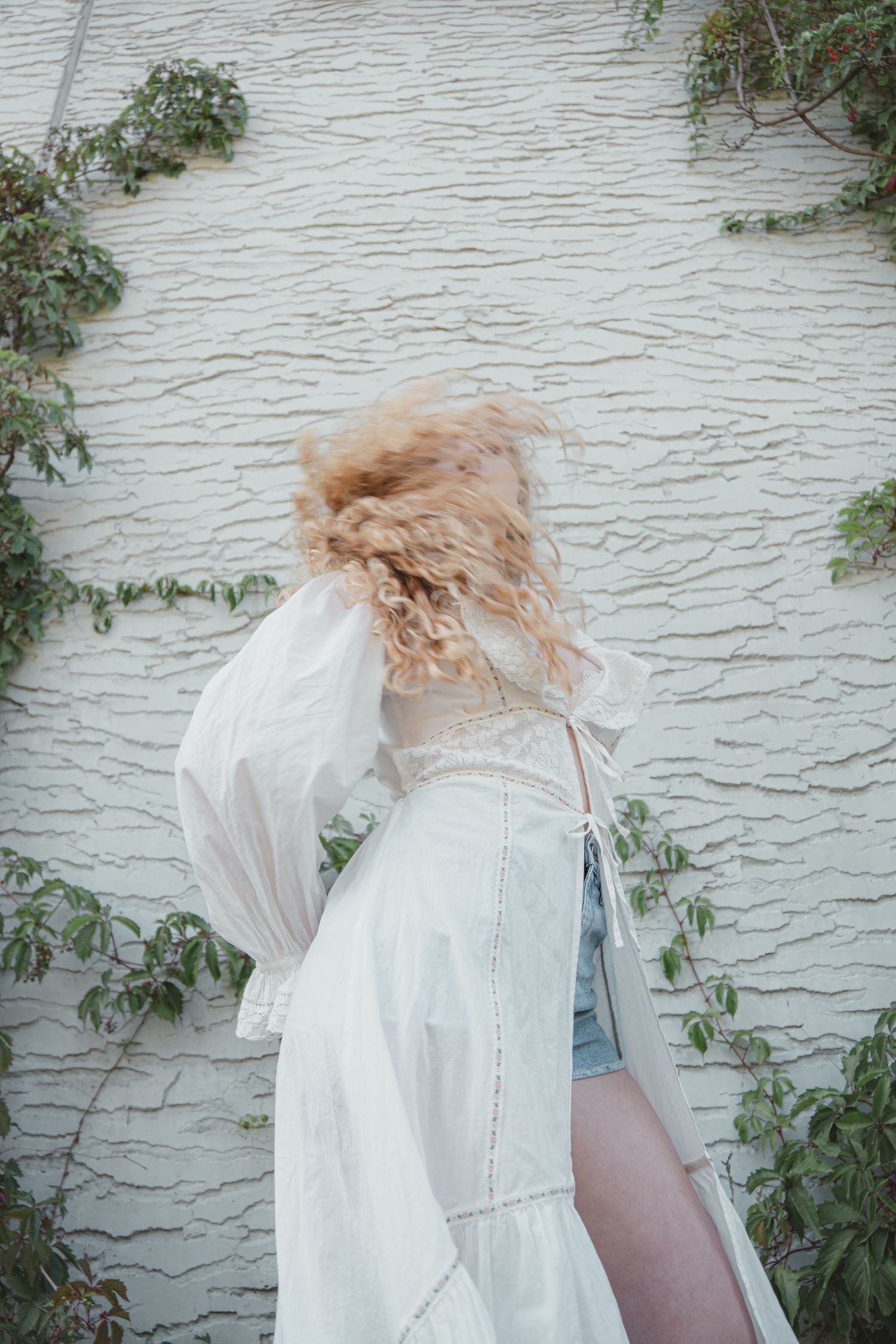 Young woman with curly blonde hair in white lace dress and denim shorts posed against ivy-covered white wall.