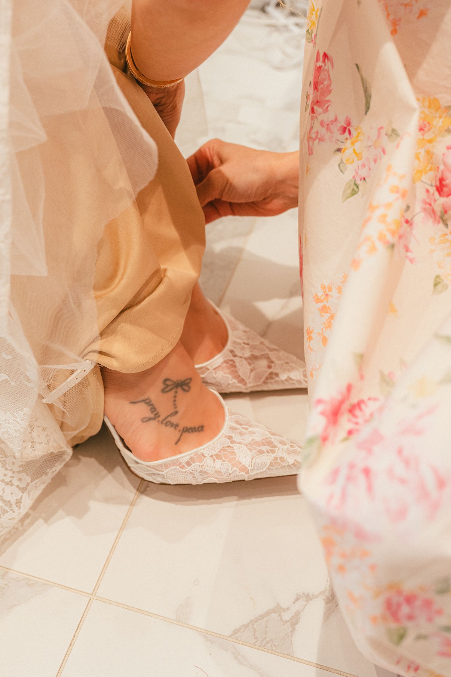 Bridesmaid helping the bride with her lace wedding shoes, revealing a delicate bow ankle tattoo.