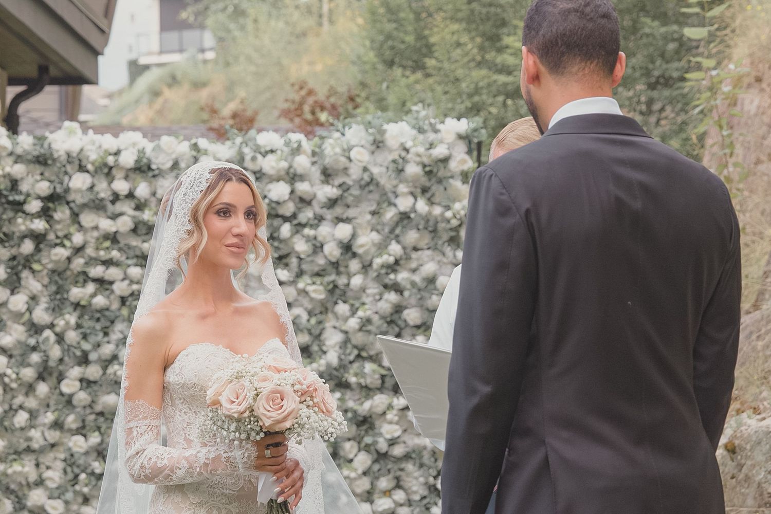 Bride in strapless gown and veil holding roses faces groom at elegant outdoor wedding ceremony.
