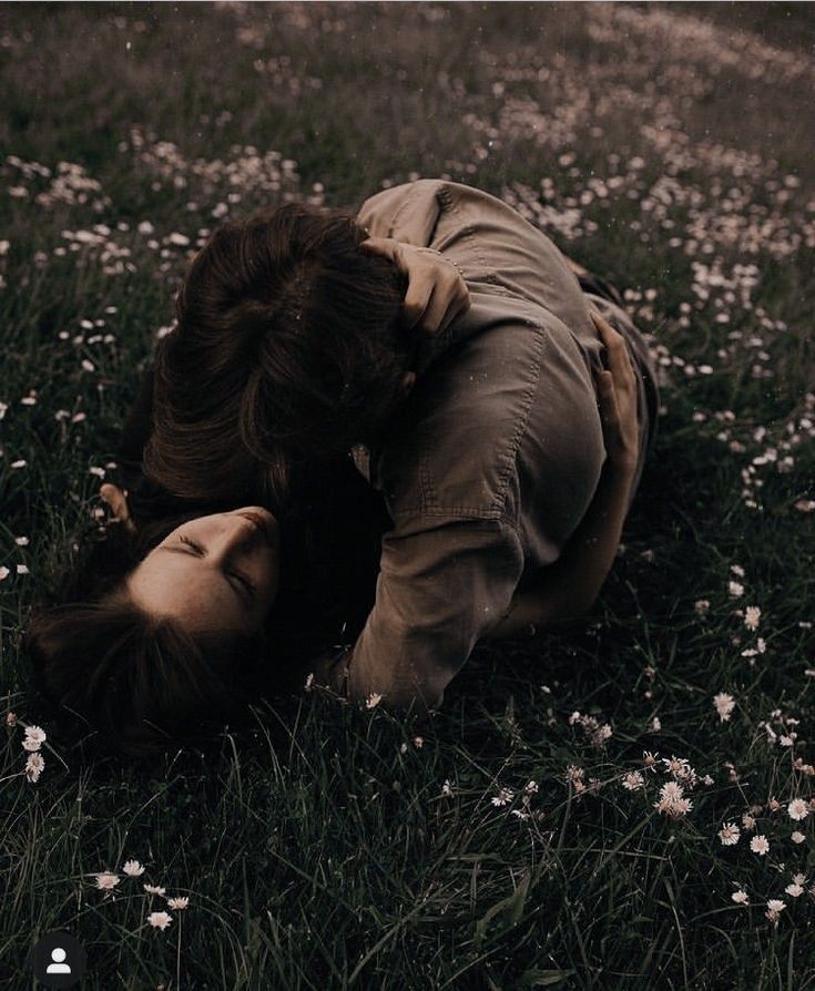 Two people lying in a flower-dotted grass field sharing an intimate embrace on a moody, romantic afternoon.