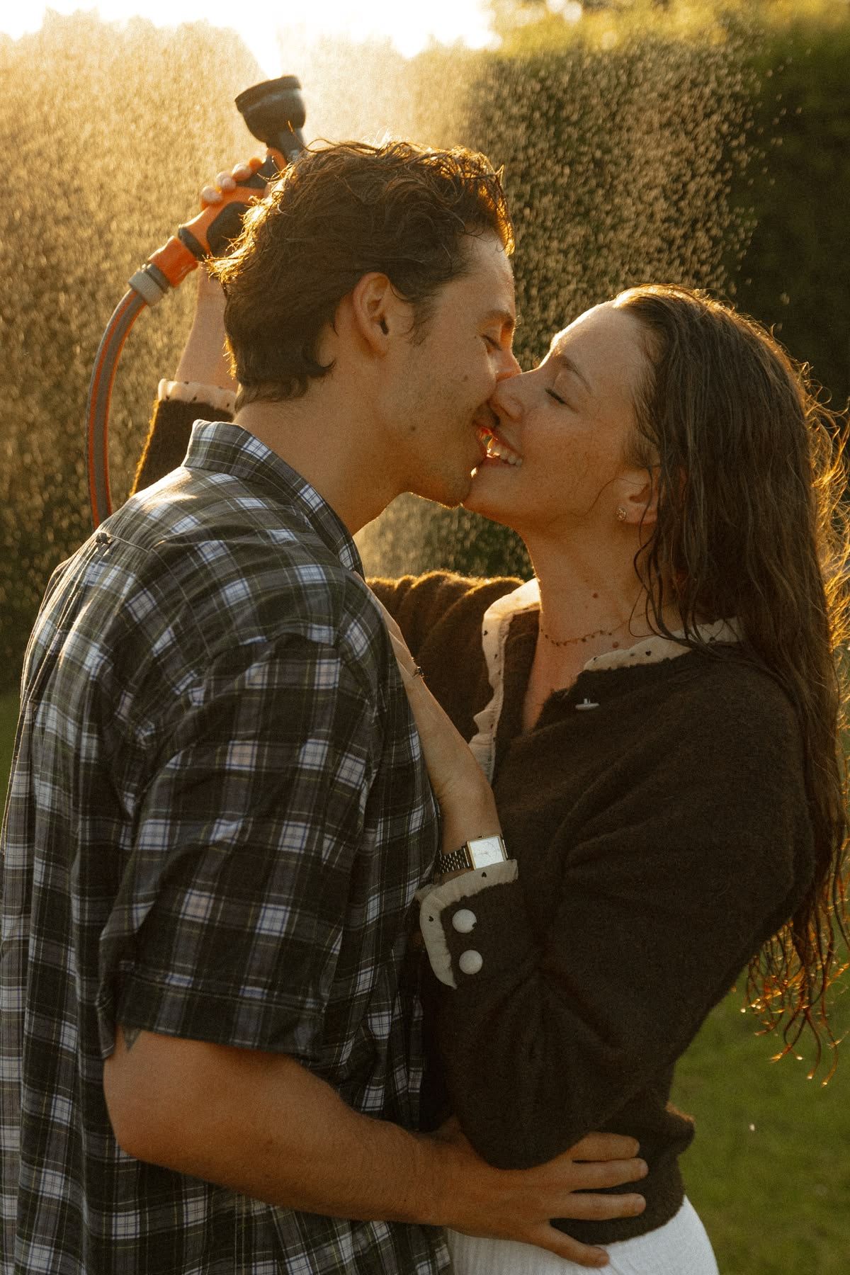 Couple kissing and laughing in garden sprinkler water spray during golden hour sunset light.