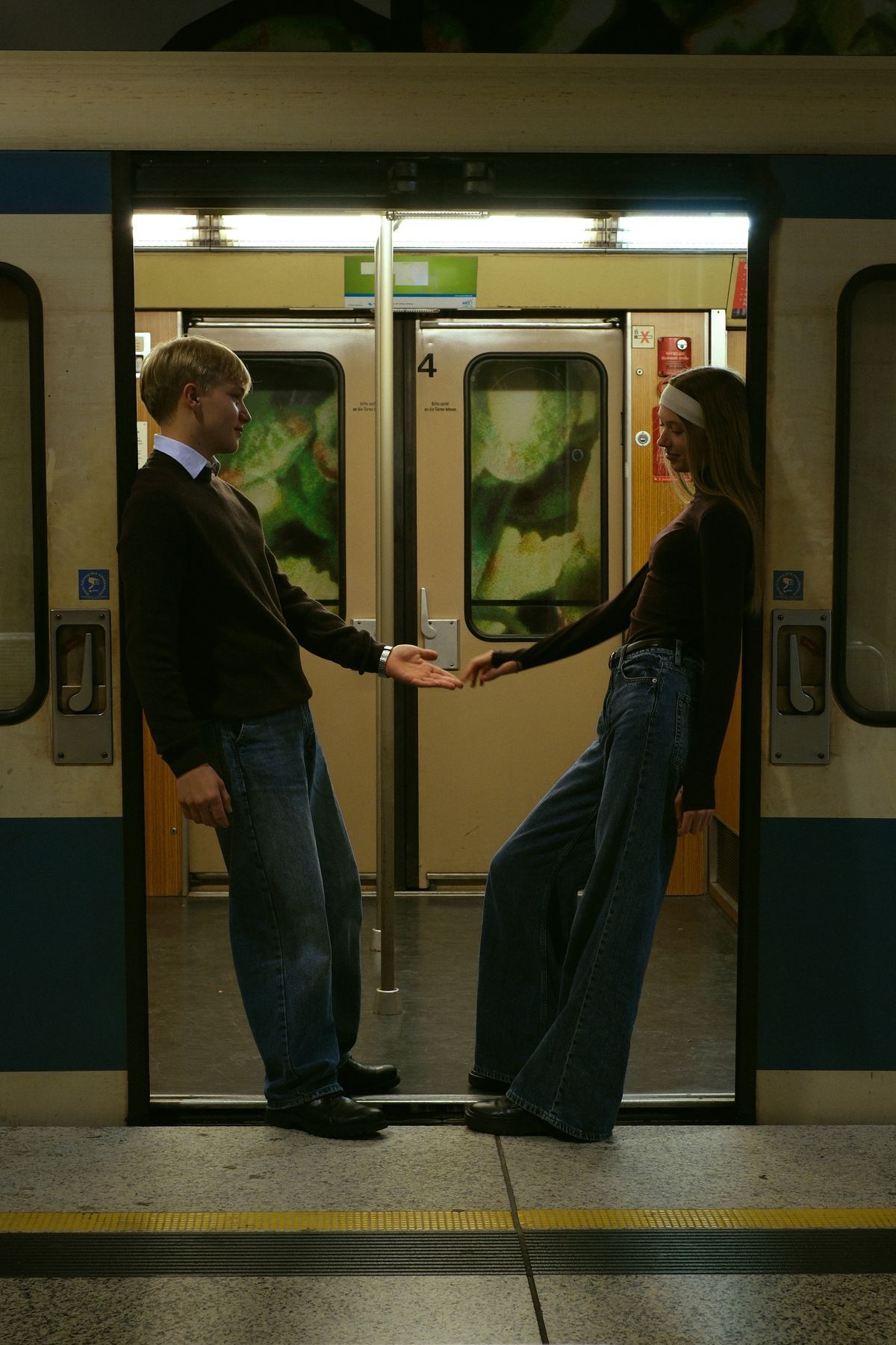 Couple holding hands in subway train doorway, facing each other on platform in jeans and dark sweaters.
