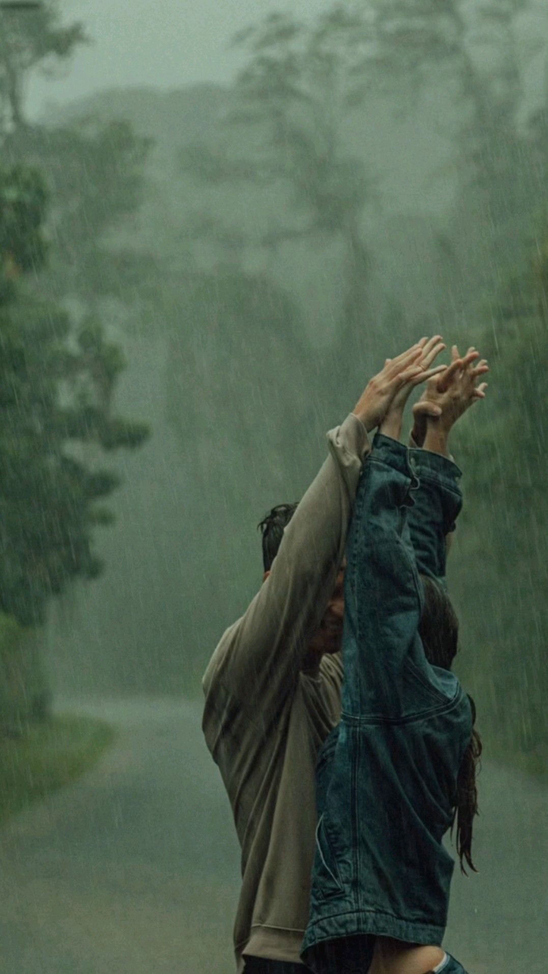 Two people dancing joyfully in heavy rain on a misty road, arms raised together toward the sky.