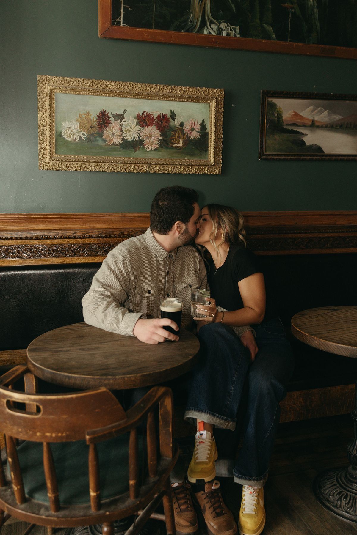 Couple sharing a kiss over dark beers in a cozy pub booth with vintage paintings on green walls.