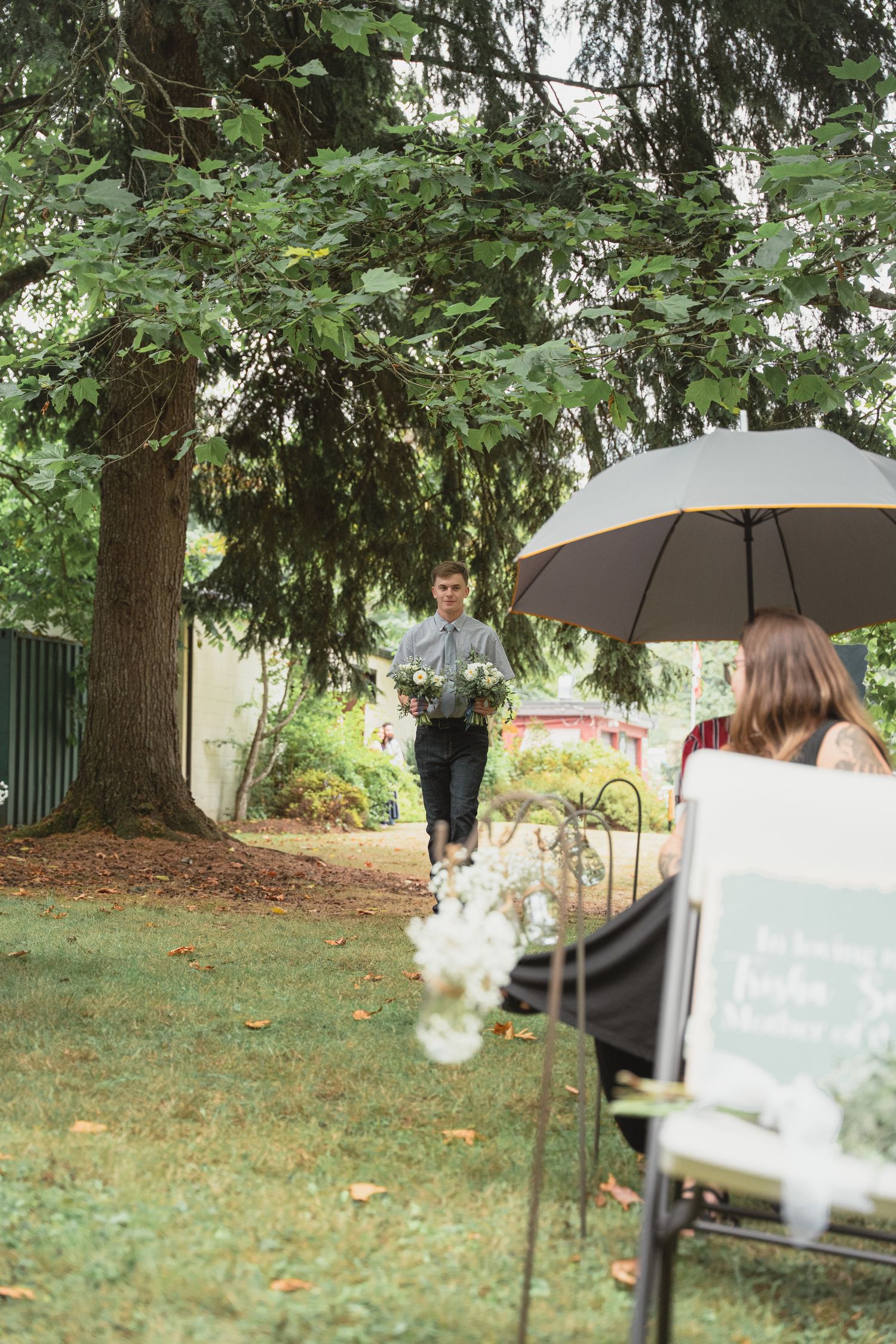 Man carrying white flowers walks toward seated guests under umbrella at outdoor wedding ceremony on green lawn.