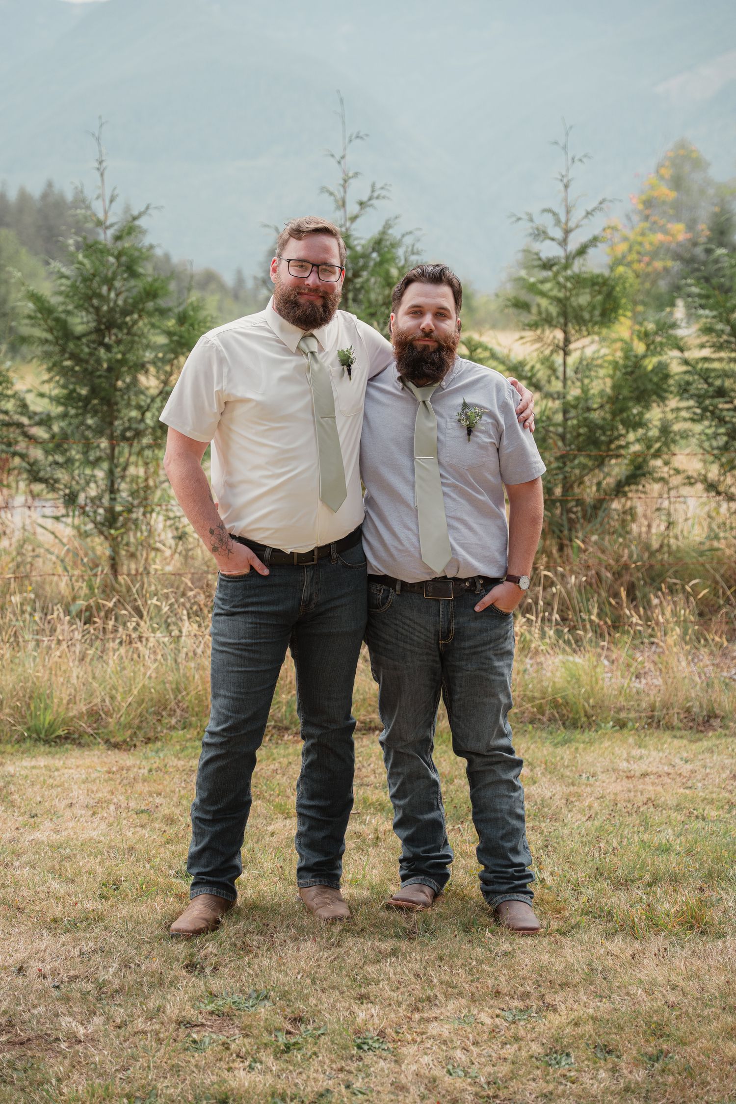 Two bearded grooms pose together smiling outdoors wearing ties and boutonnieres on their wedding day.