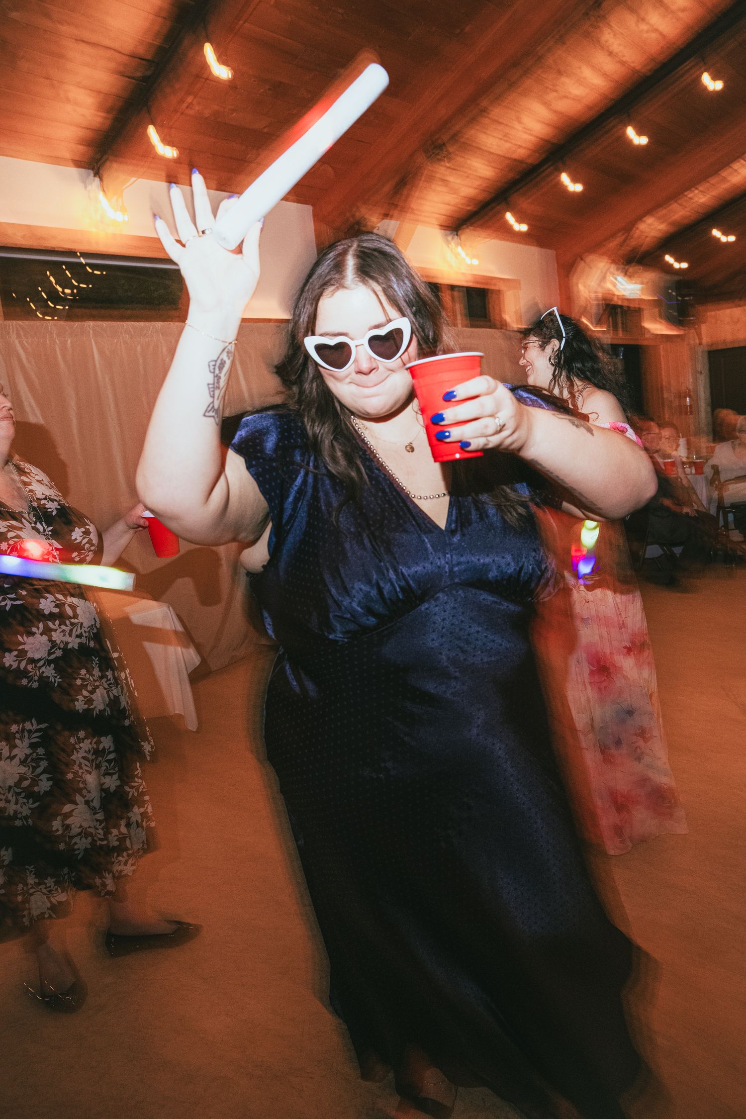 Woman in navy dress dancing with glow stick and red solo cup at wedding reception.